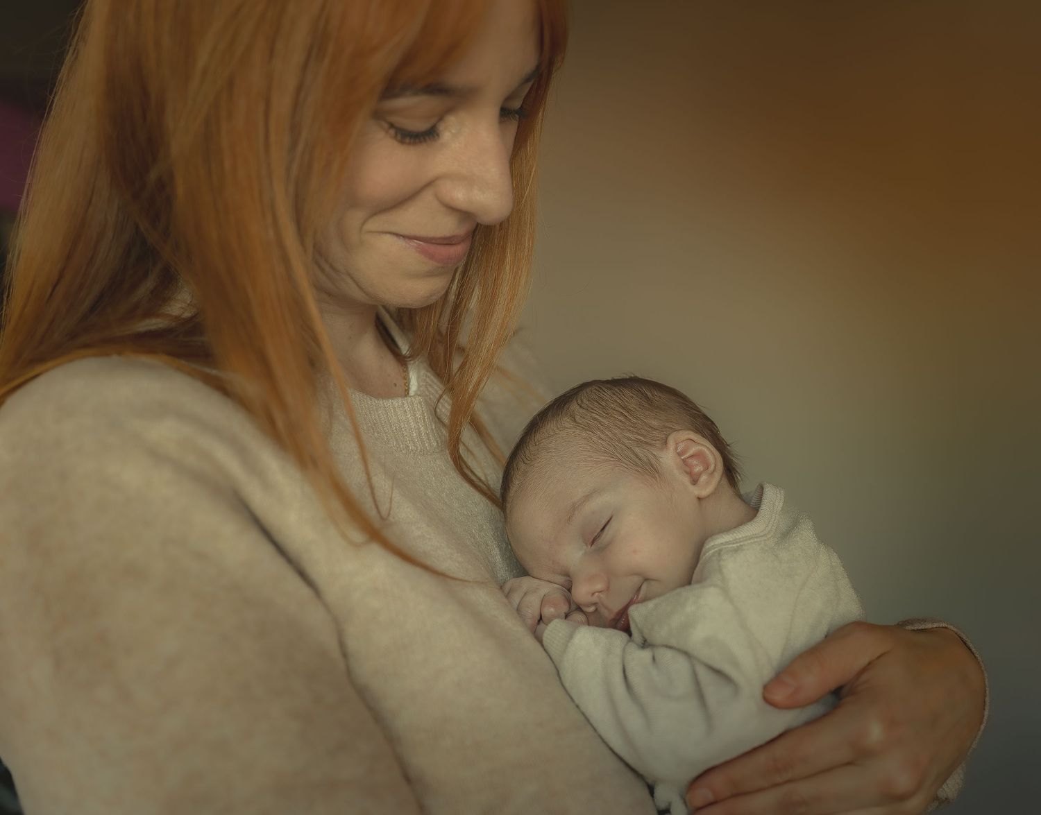 A red-haired mother tenderly holds her sleeping newborn baby in her arms, smiling softly.