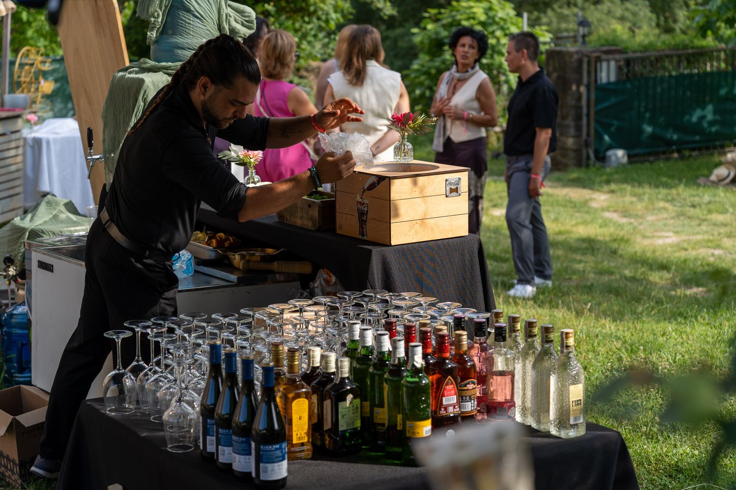 Bartender preparing drinks at an outdoor bar setup with rows of wine bottles and glasses at a garden party event.