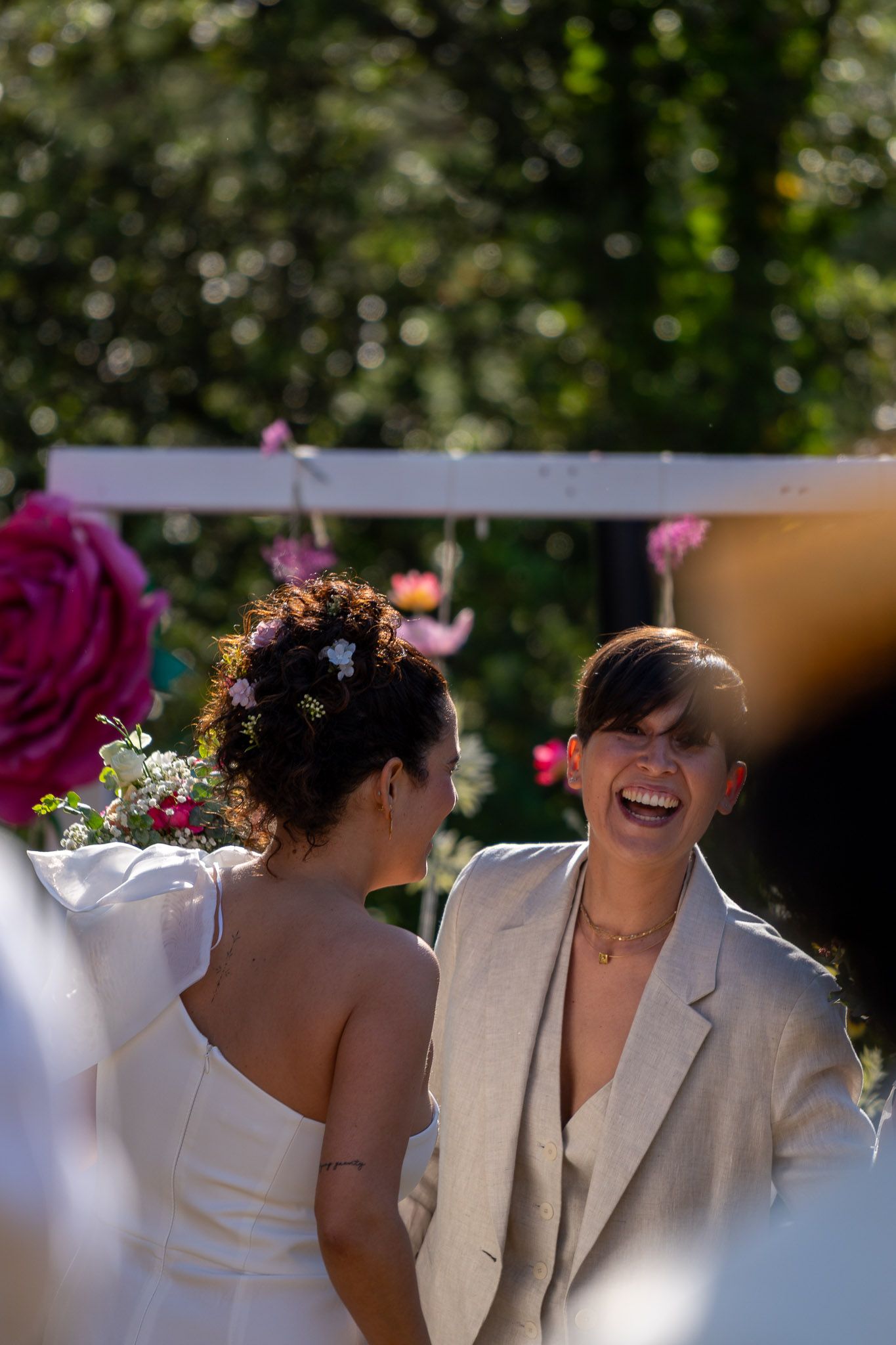 Two brides laughing joyfully at outdoor wedding ceremony surrounded by pink roses and lush greenery.