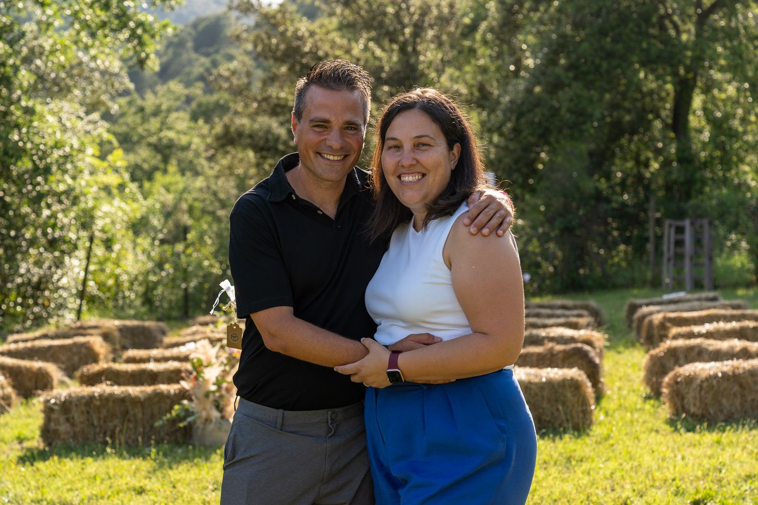 Smiling couple embracing outdoors on green grass with hay bales and trees in background on a sunny day.