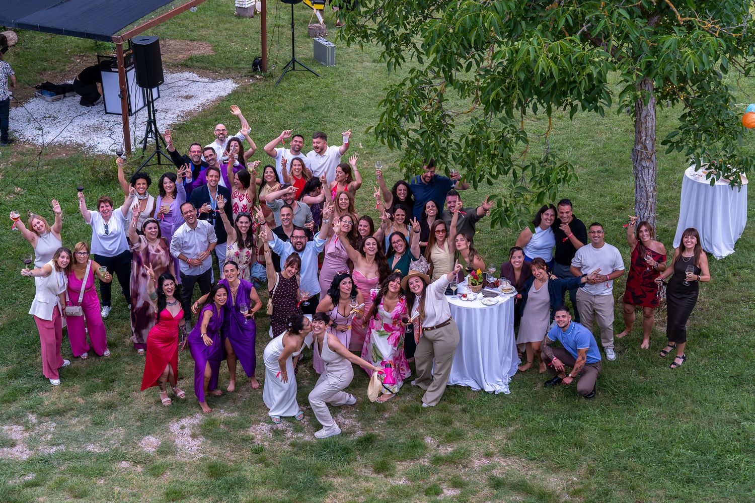 Large group of joyful wedding guests posing outdoors on green lawn under tree at elegant summer celebration.