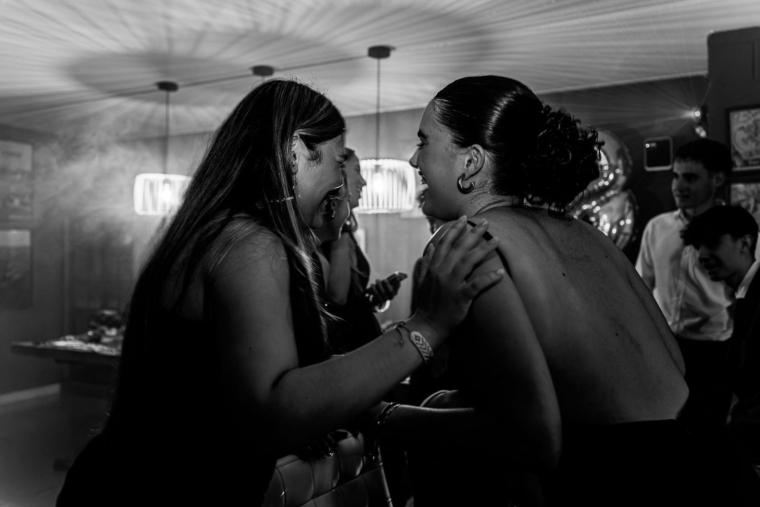 Two women share an intimate conversation at a dimly lit bar in this black and white candid photo.