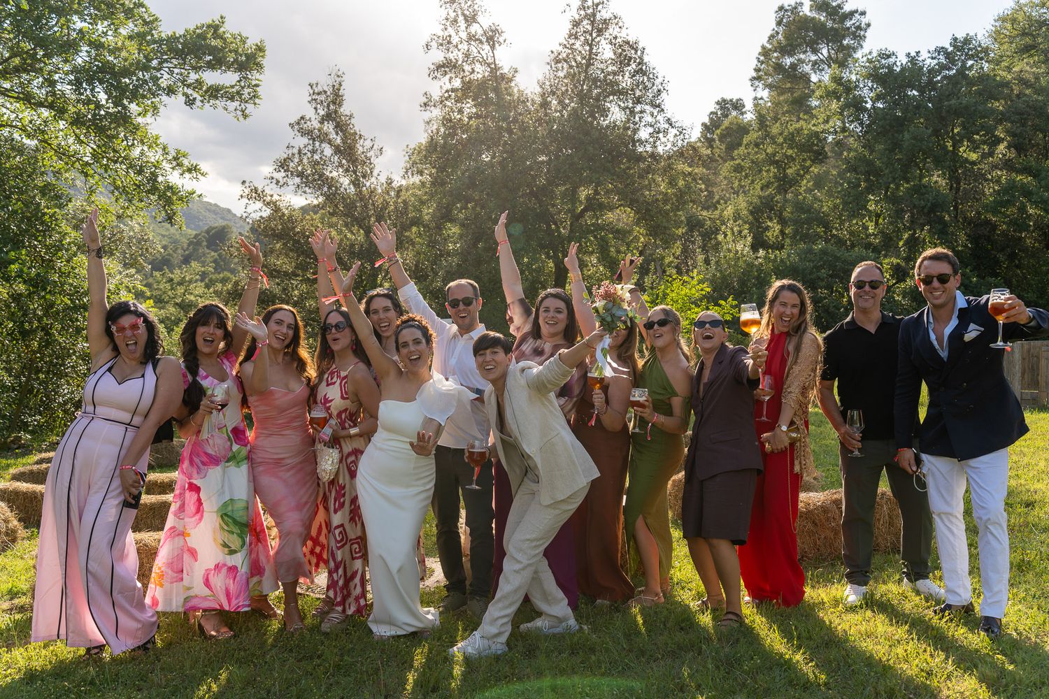 Joyful wedding party group photo outdoors, guests in colorful attire celebrating with raised arms in sunlit garden.