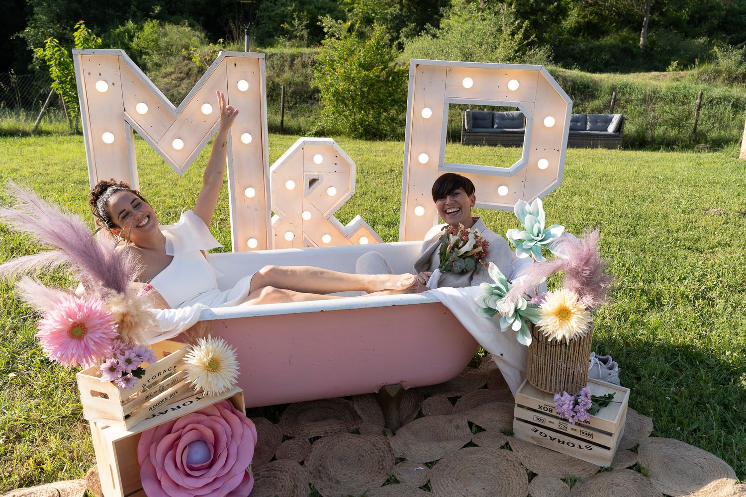 Couple posing in a pink bathtub outdoors with marquee letters M&P, surrounded by flowers and fairy lights.