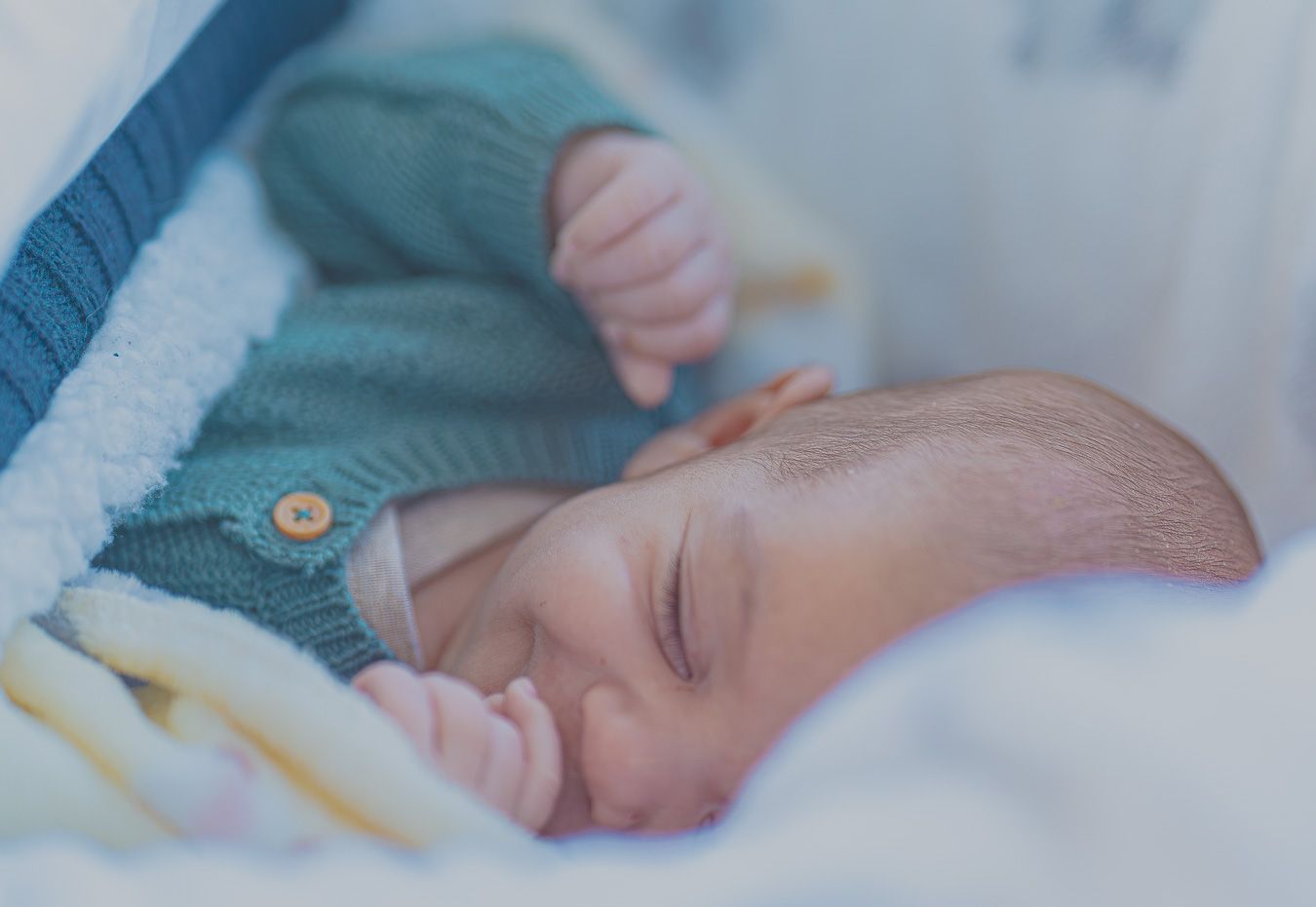 Peaceful newborn baby sleeping in a white blanket wearing a teal knit onesie.