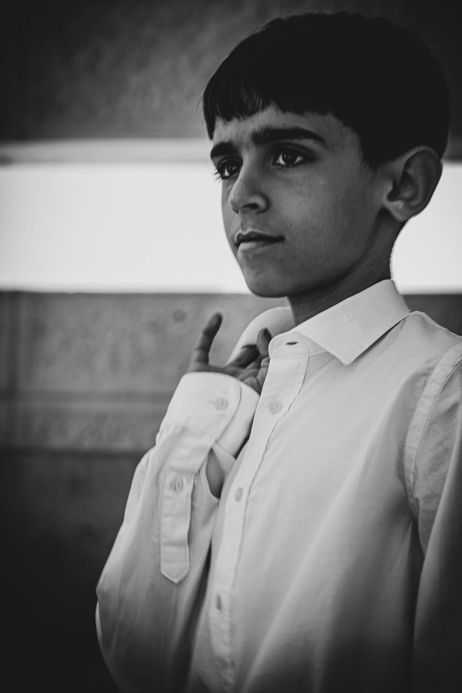 Black and white portrait of a young boy in a white dress shirt, adjusting his collar with a thoughtful expression.