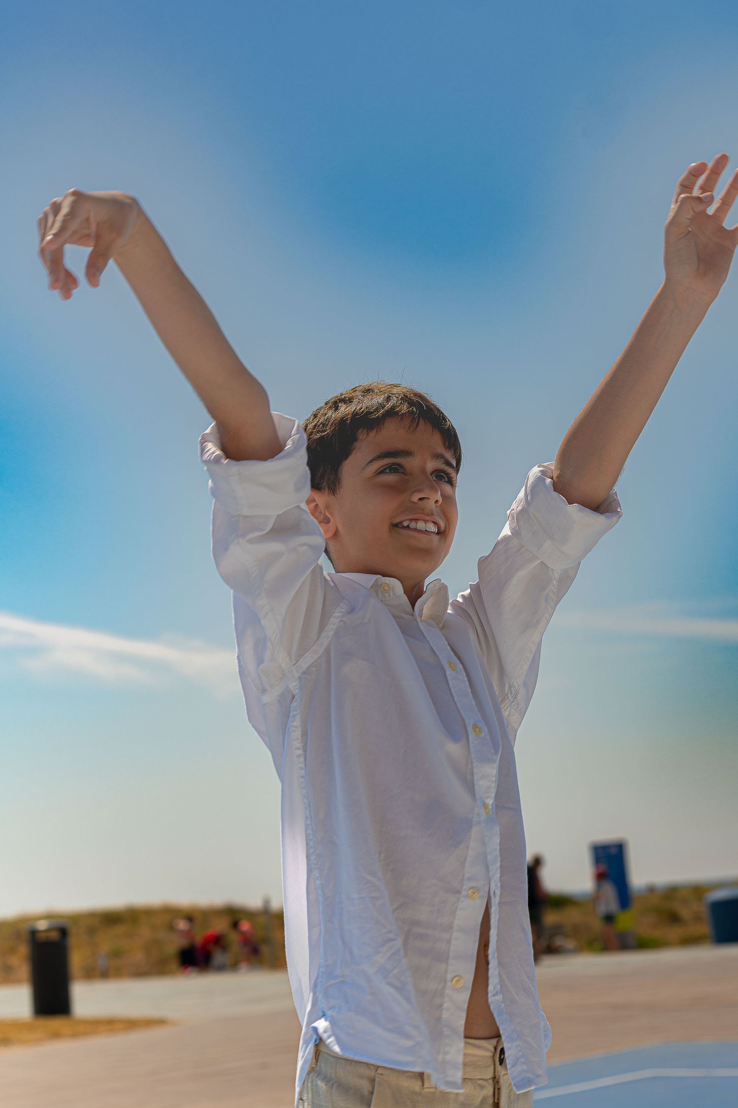 A joyful boy in a white shirt raises his arms triumphantly under a bright blue sky outdoors.