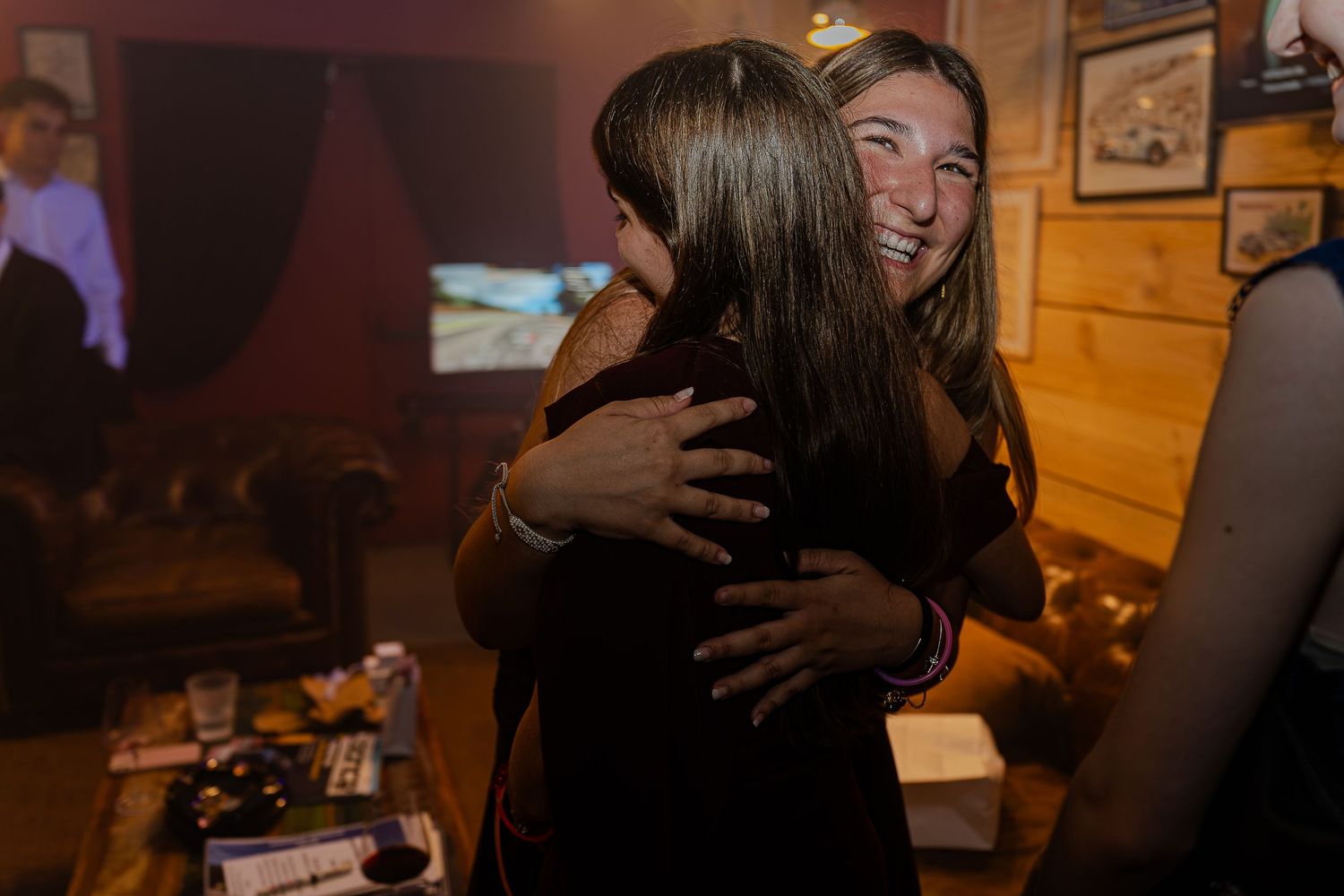Two women sharing a warm embrace and smiling at an indoor party in a cozy wooden venue.