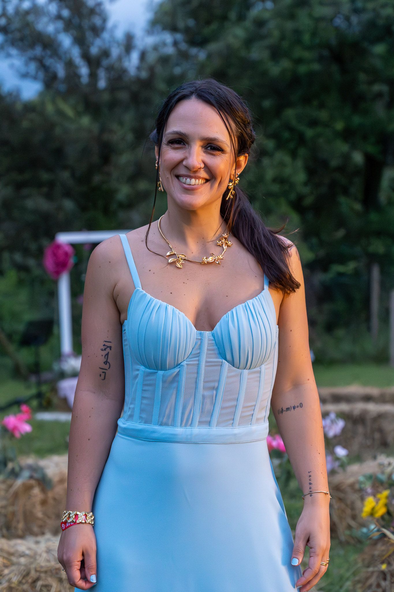 Smiling woman in light blue dress with gold jewelry and tattoos stands outdoors near flowers at dusk.