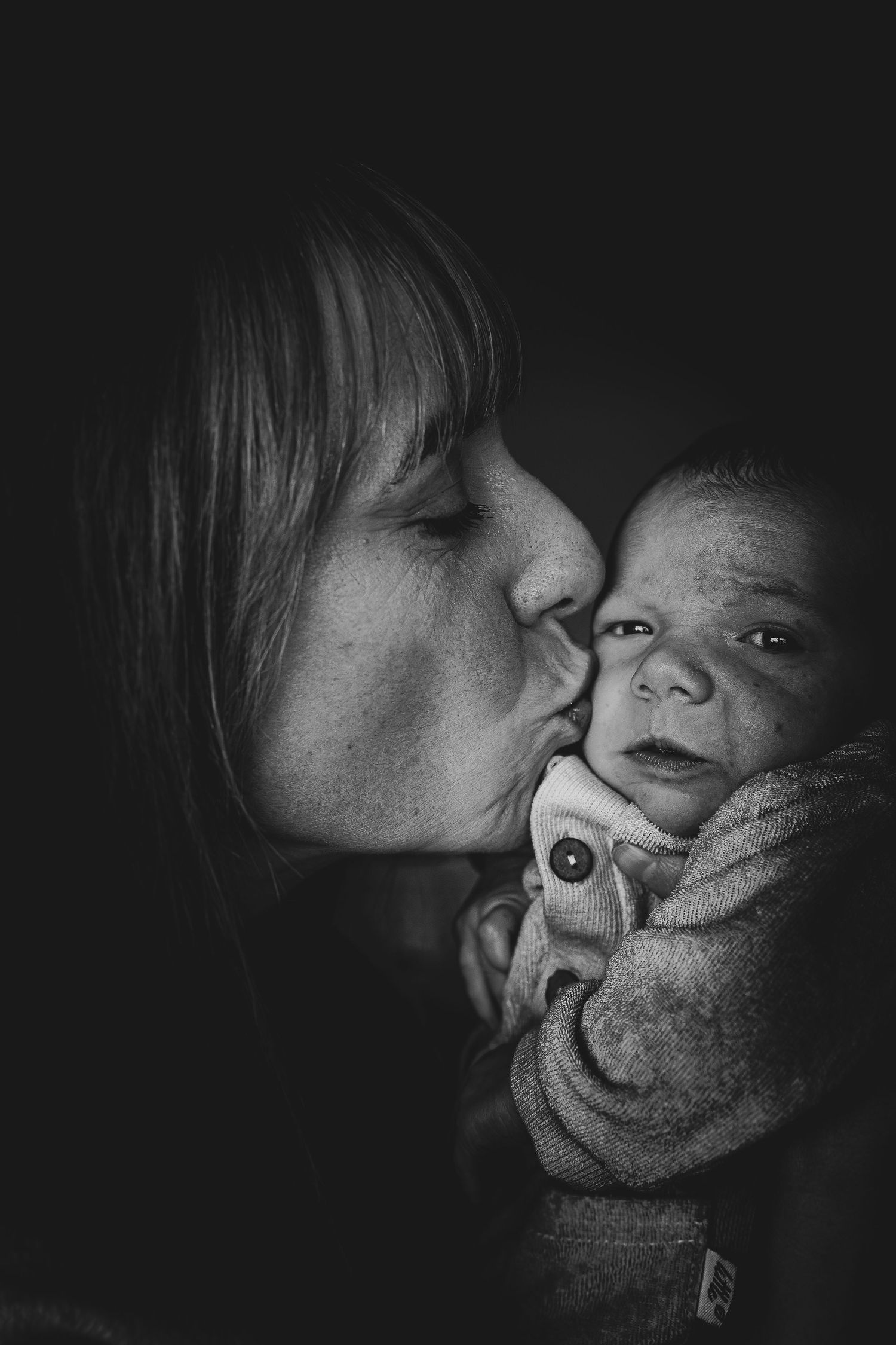 Foto en blanco y negro de madre besando a su bebé recién nacido. Reportaje newborn documental y fotografía familiar en Barcelona.