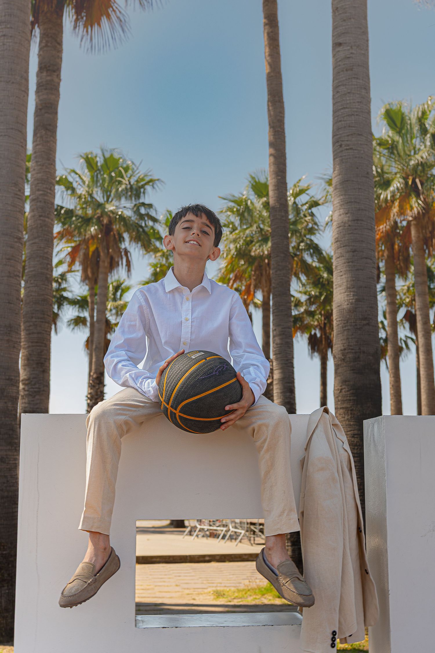 A young boy in white shirt holds basketball while sitting on white structure with tall palm trees in background.