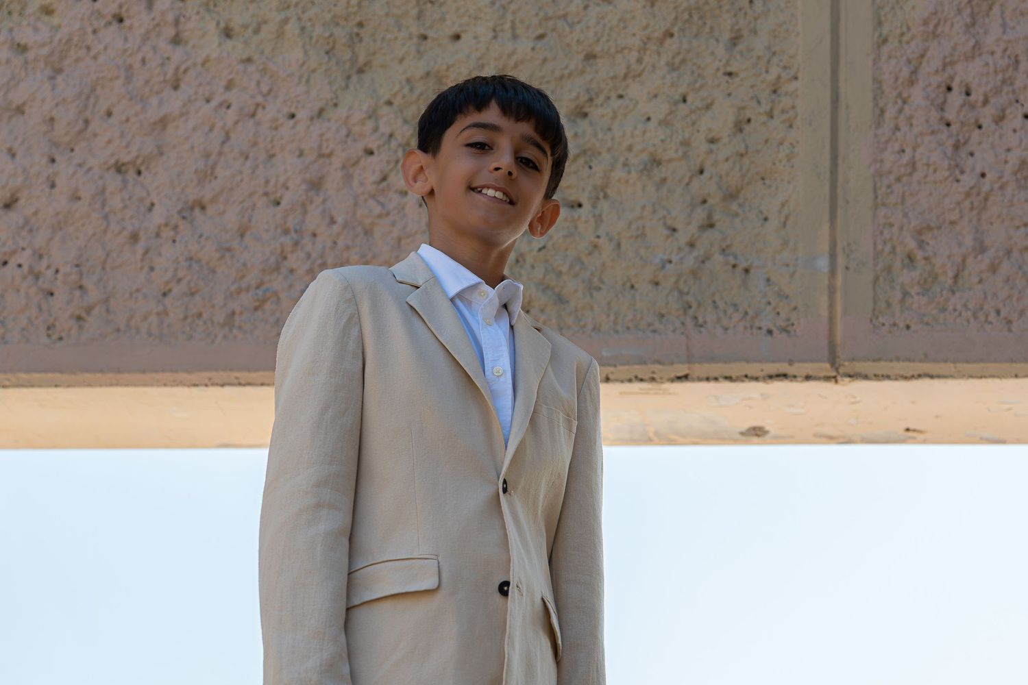 A young boy smiling in a beige suit and blue tie standing outdoors.