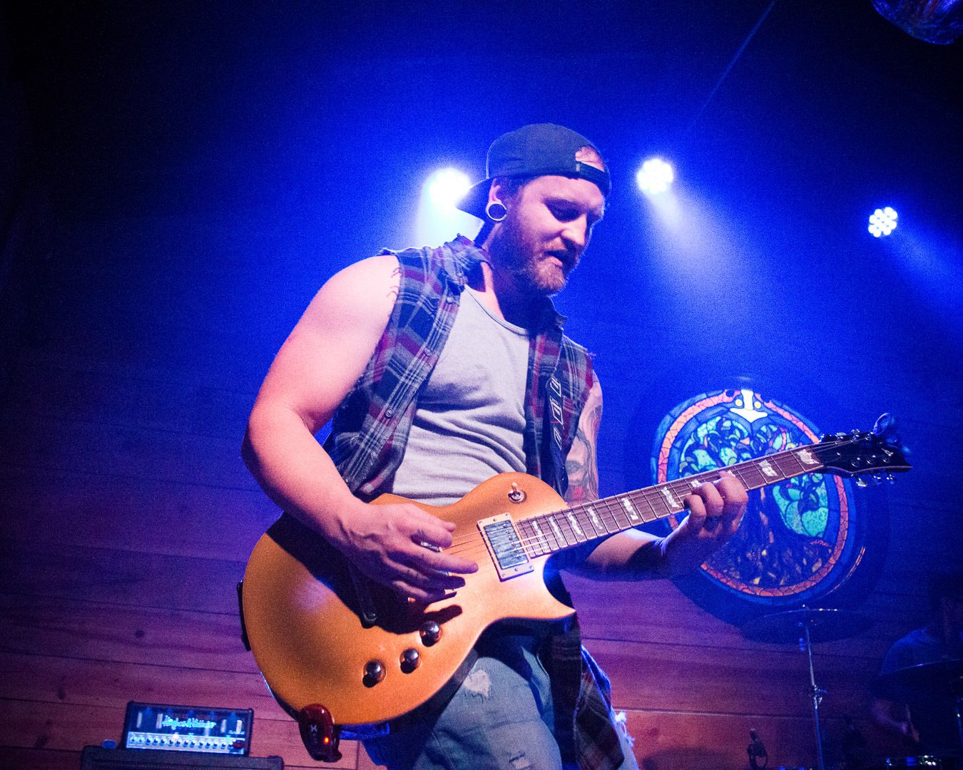 Male guitarist performing live on stage under blue and purple lights, wearing a backward cap and plaid shirt, concert photography.