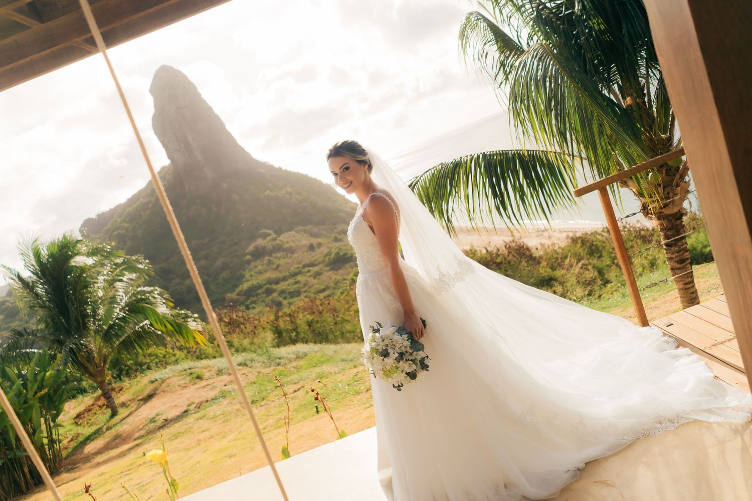 A bride in a white wedding dress poses against a backdrop of palm trees and mountains in a tropical setting.