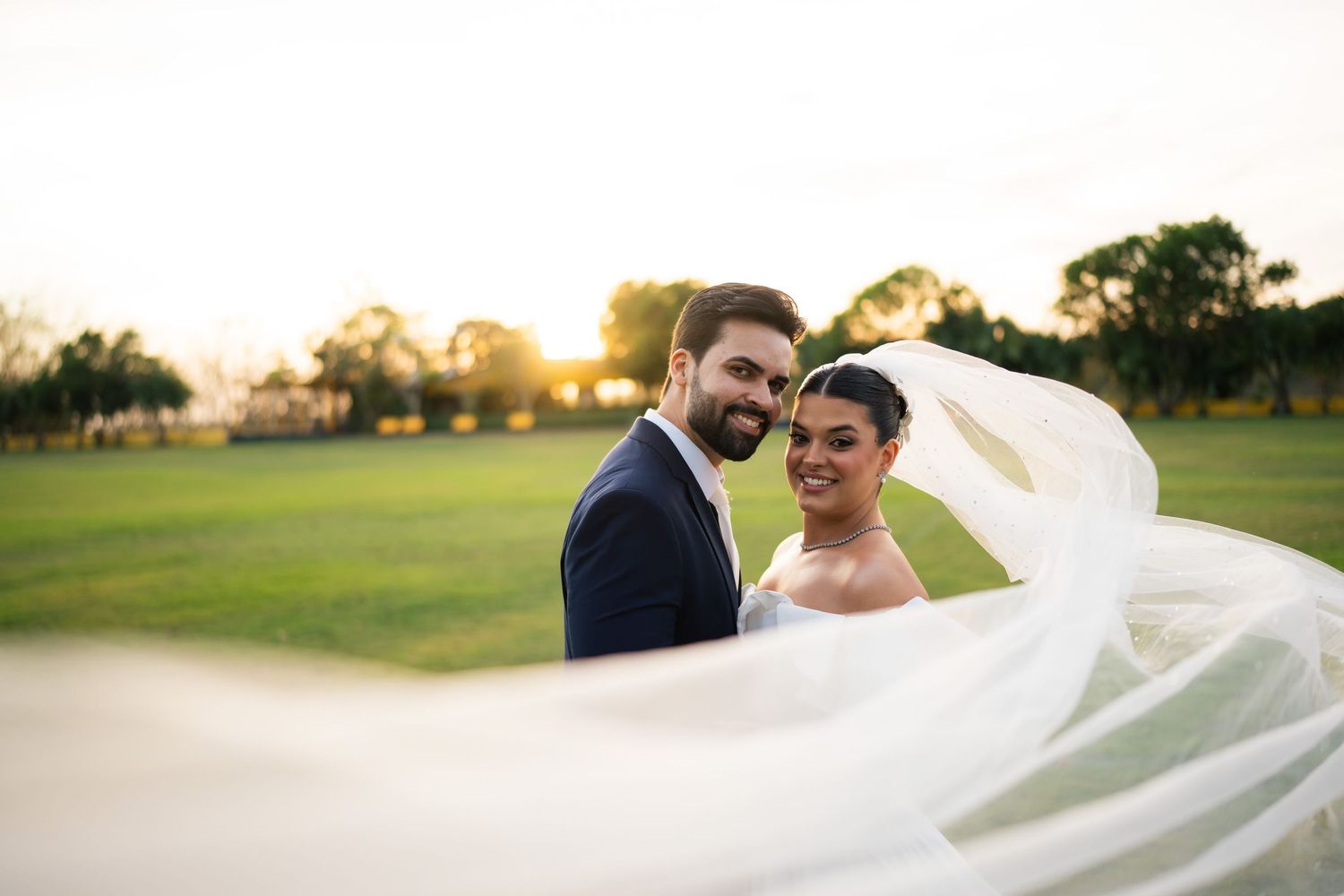 Romantic wedding couple embracing in a sunlit outdoor setting with a flowing cathedral veil catching the breeze.