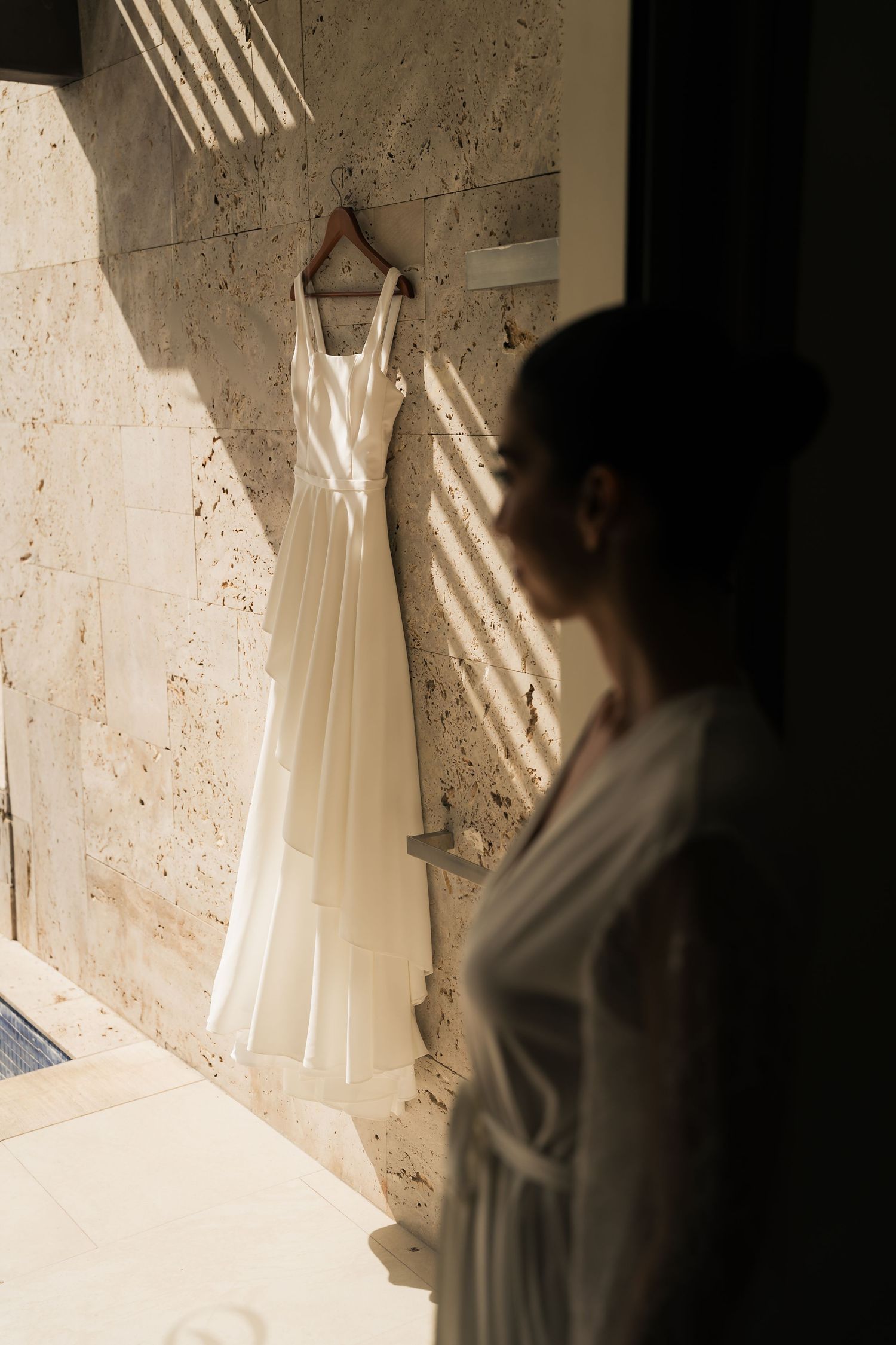 A white wedding dress hangs elegantly in a sunlit room casting dramatic shadows on a textured wall.