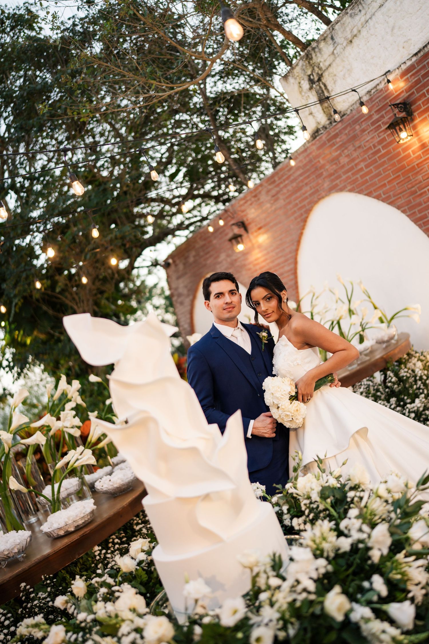Elegant wedding portrait among white flowers and string lights against a brick wall.