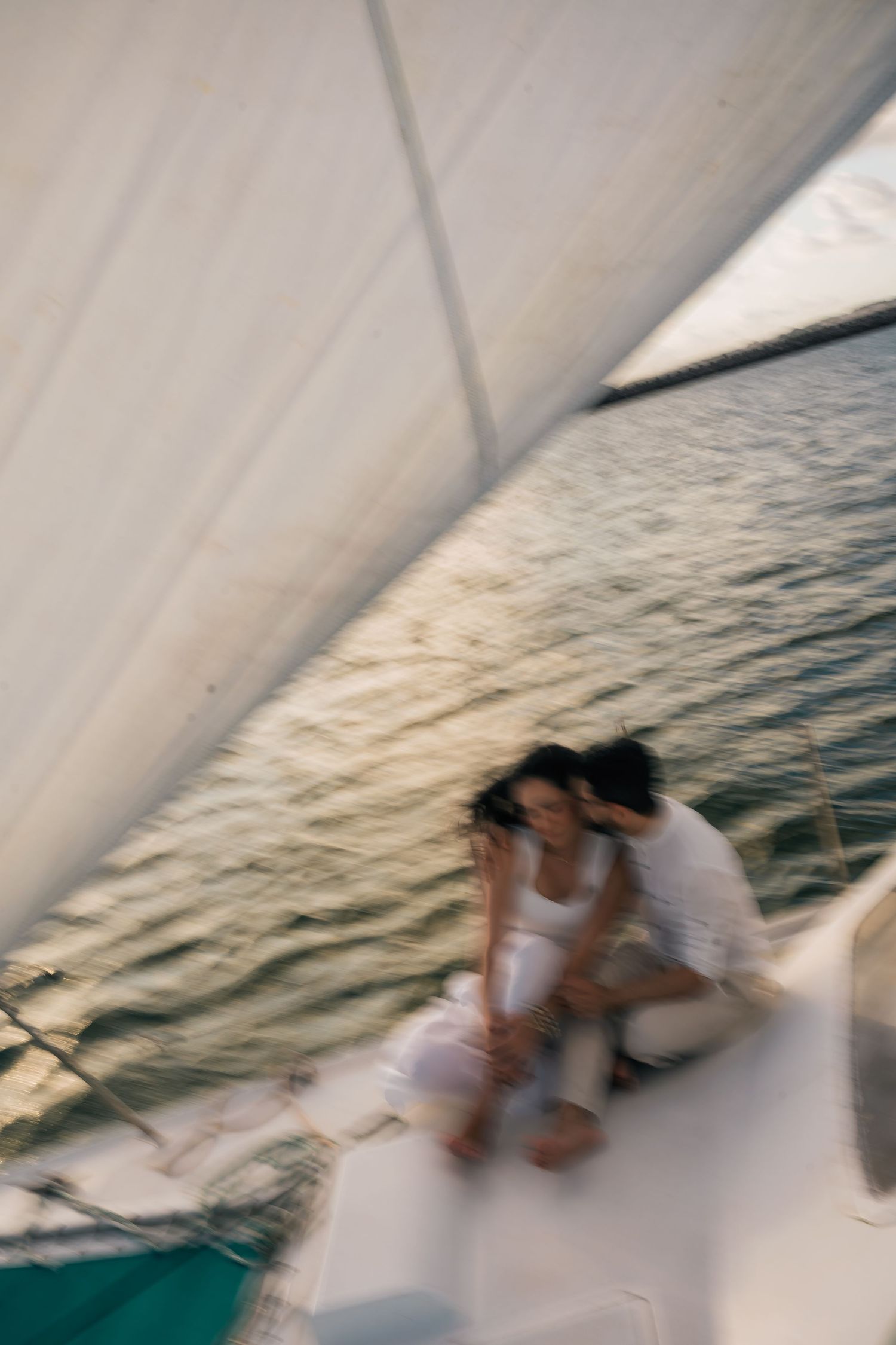 Motion-blurred view of a sailboat deck with white sails and ocean waves in the background at sunset.