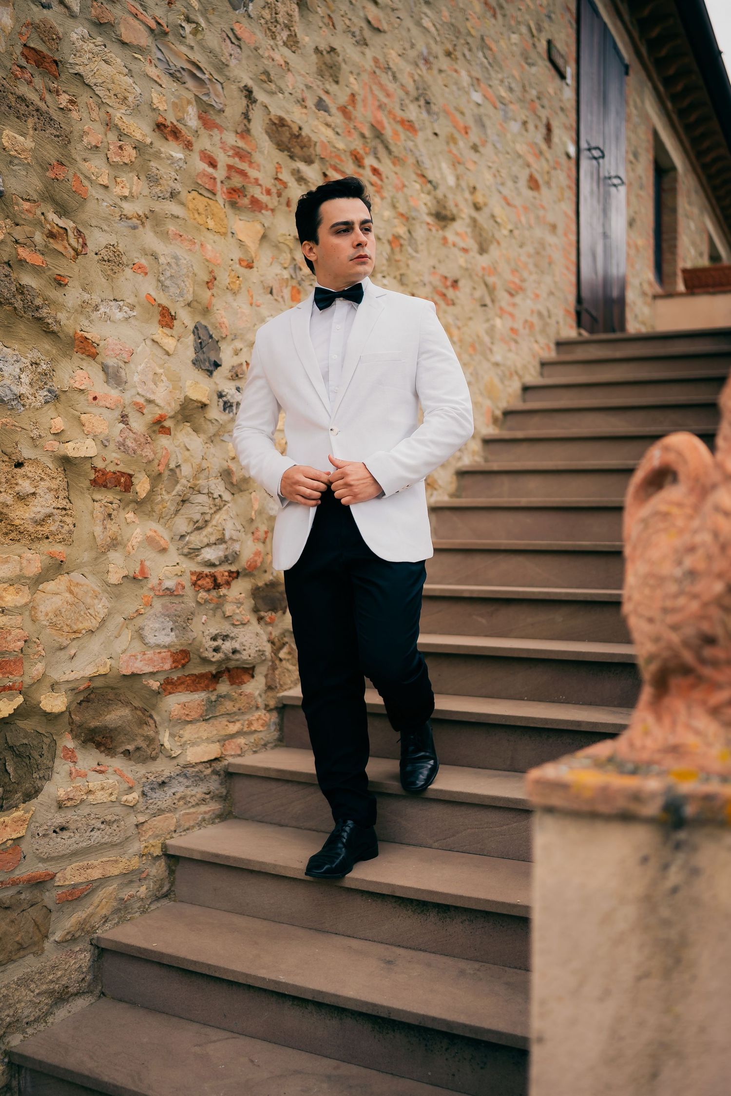 A formal groom in a white shirt and black bow tie sits on stone steps against a rustic brick wall.