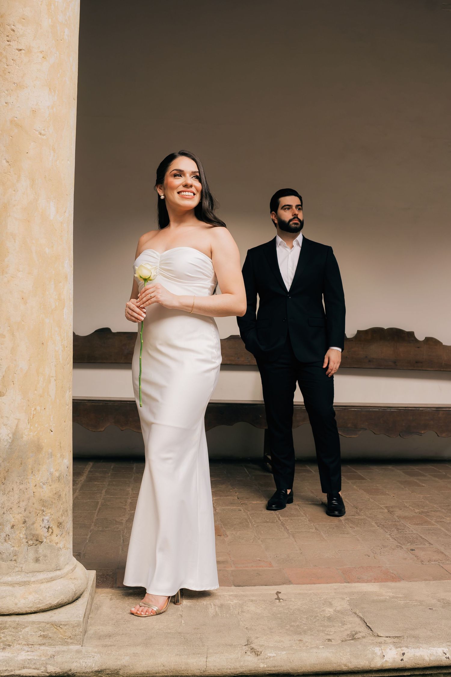 A bride in a white dress and groom in a black suit pose together between classic stone columns.