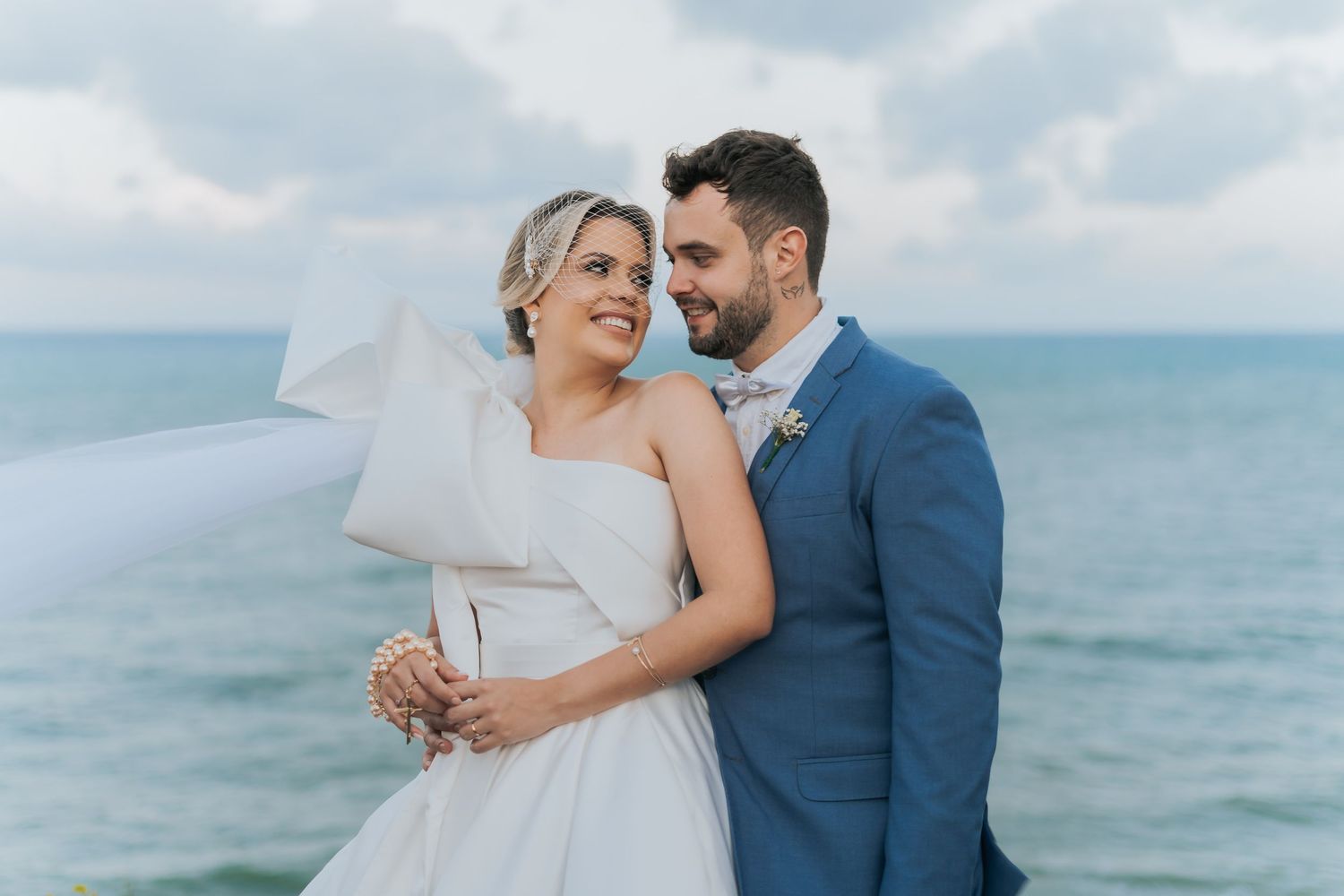 Bride and groom embrace smiling by ocean, bride in white dress with veil blowing, groom in blue suit on wedding day.