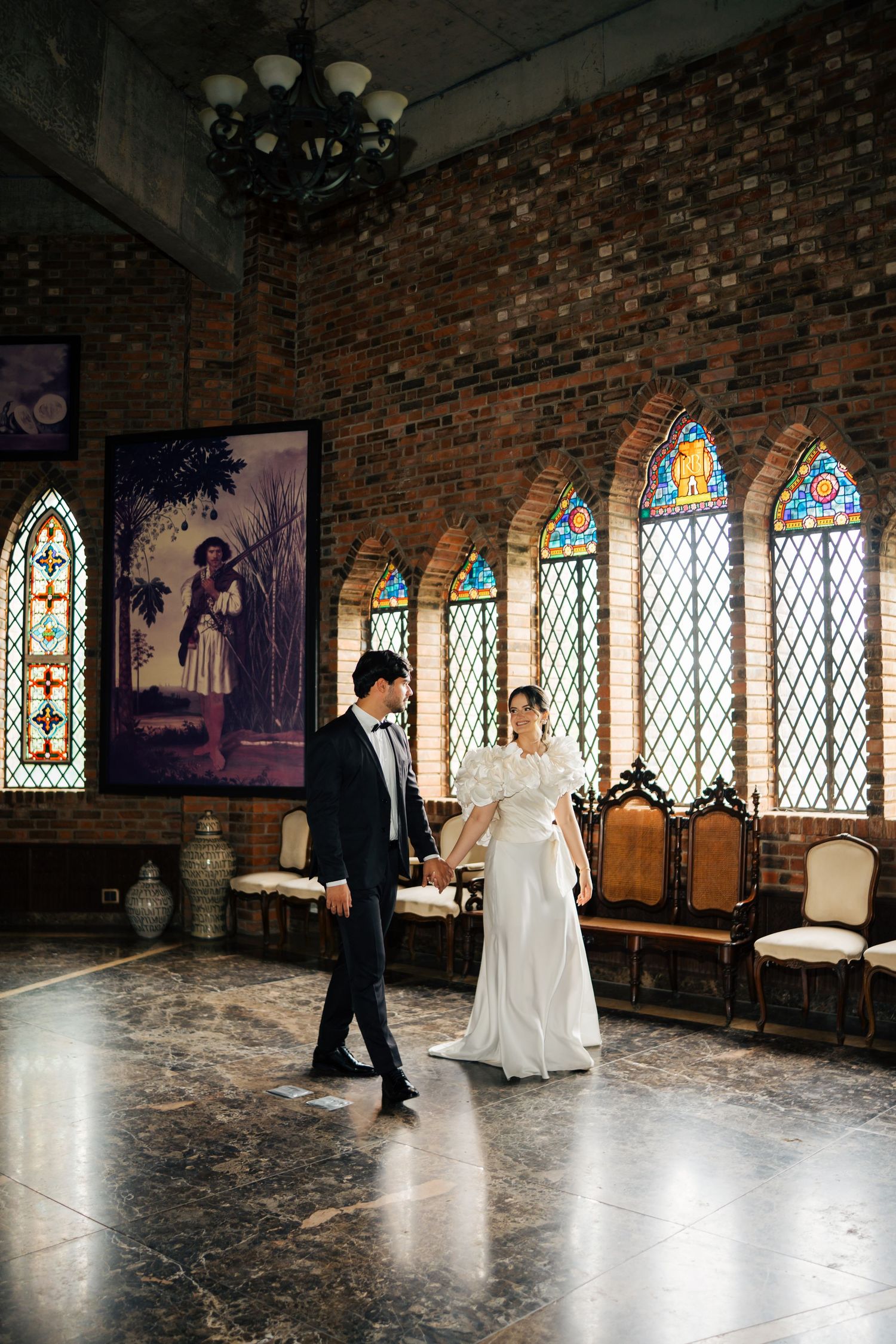 A couple dances in an elegant church hall with stained glass windows and exposed brick walls.