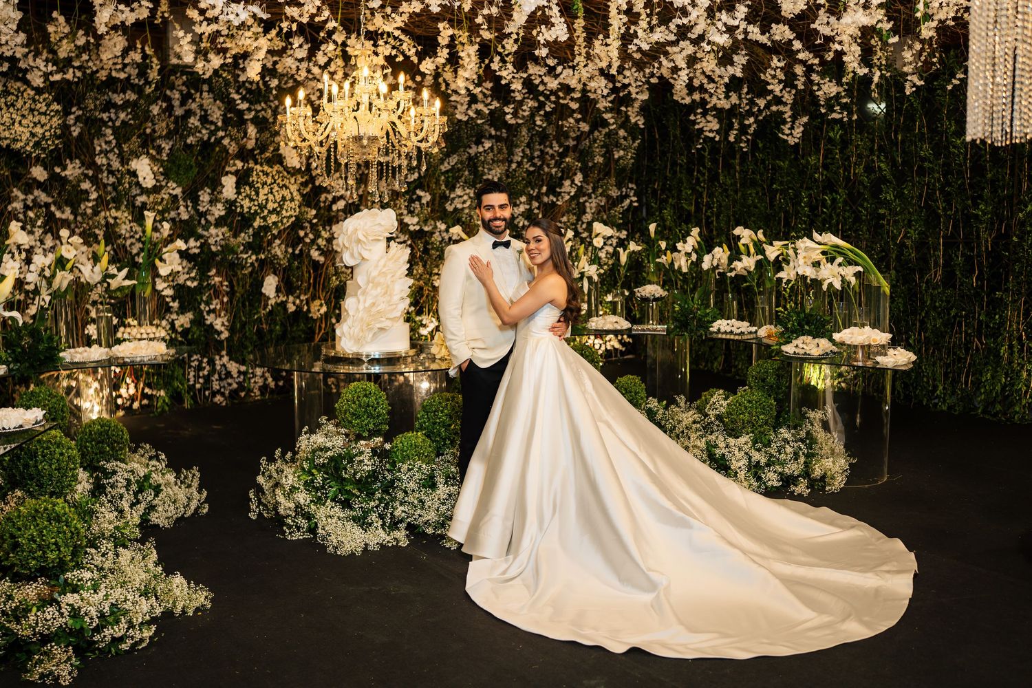 Wedding couple poses under crystal chandelier with floral decor and elegant lighting.