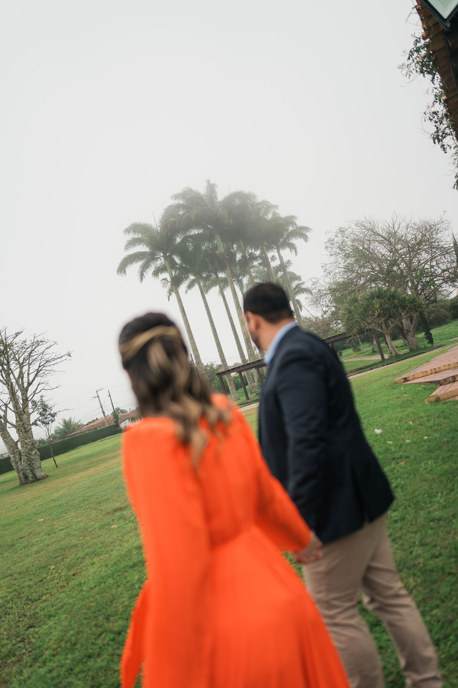 A couple walks through a palm-lined garden path, with one wearing a vibrant orange dress.