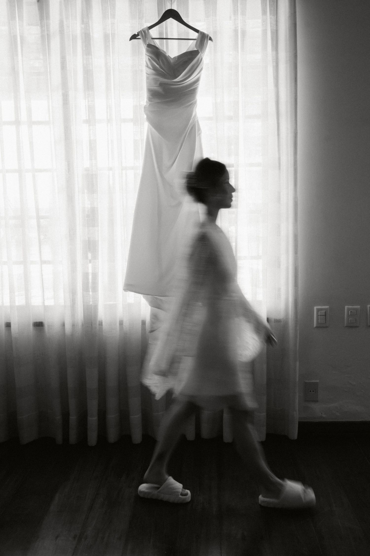 A silhouetted figure walks past an elegant white wedding dress hanging by a sunlit window in a black and white photo.