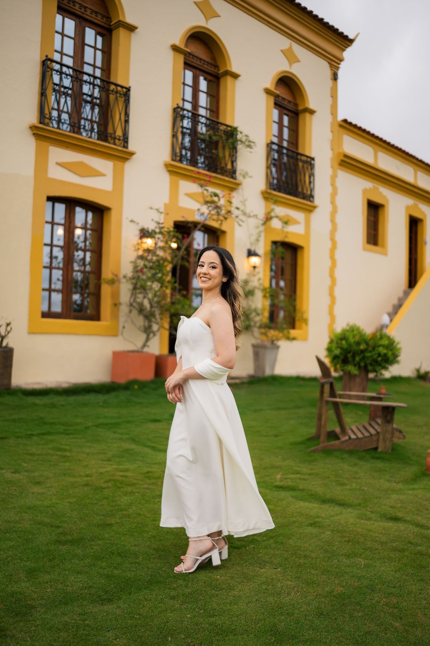 A person in a white flowing dress stands on a manicured lawn in front of a yellow Spanish colonial style building.