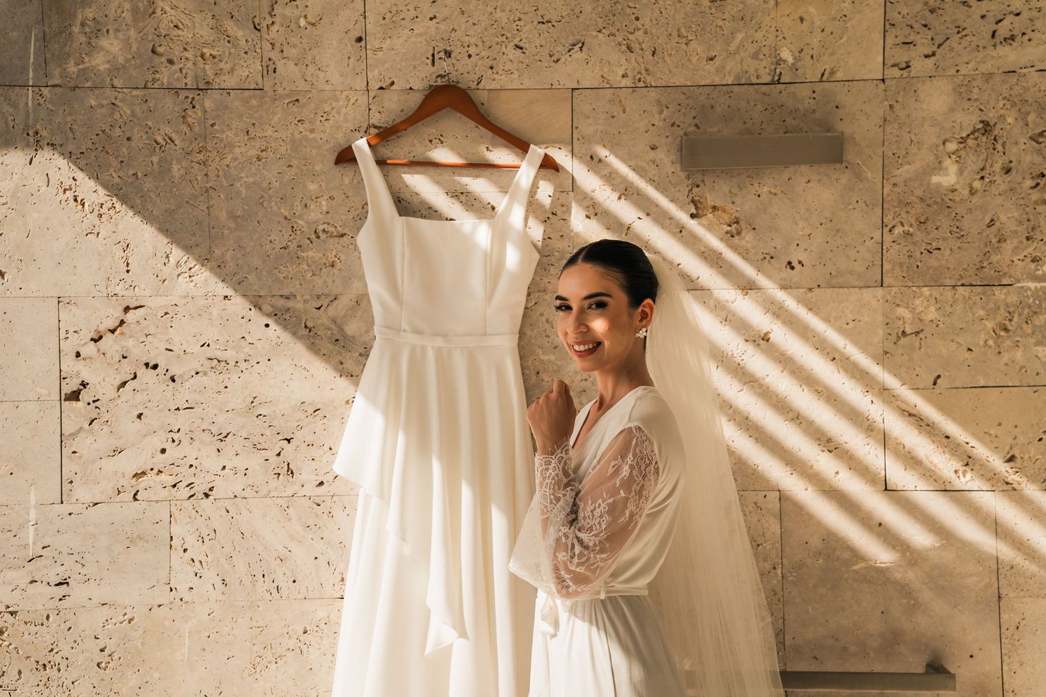 A white wedding dress hangs on a wooden hanger against a textured stone wall with dramatic natural lighting.