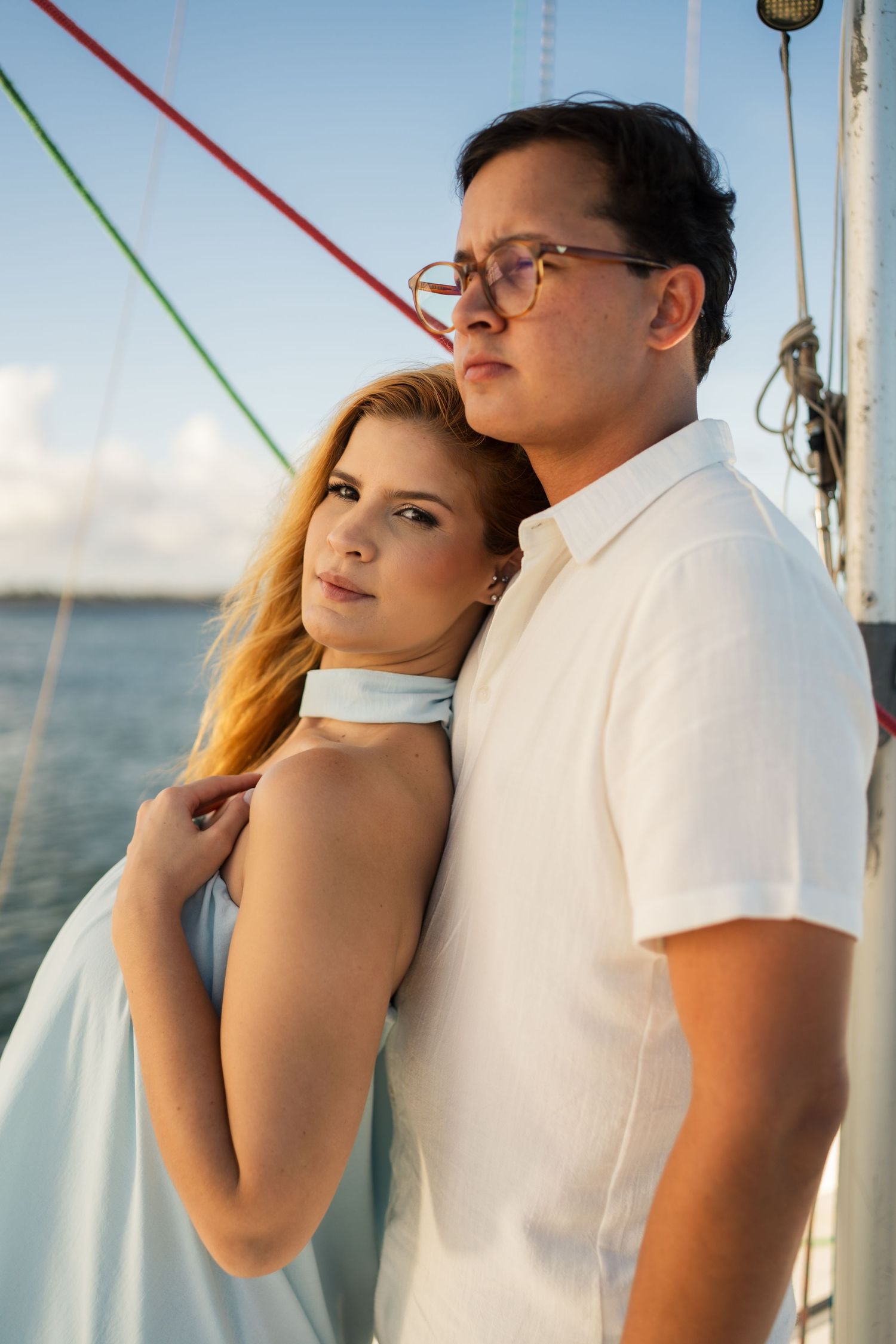 Romantic couple poses intimately against sailboat rigging with ocean and sky in background.
