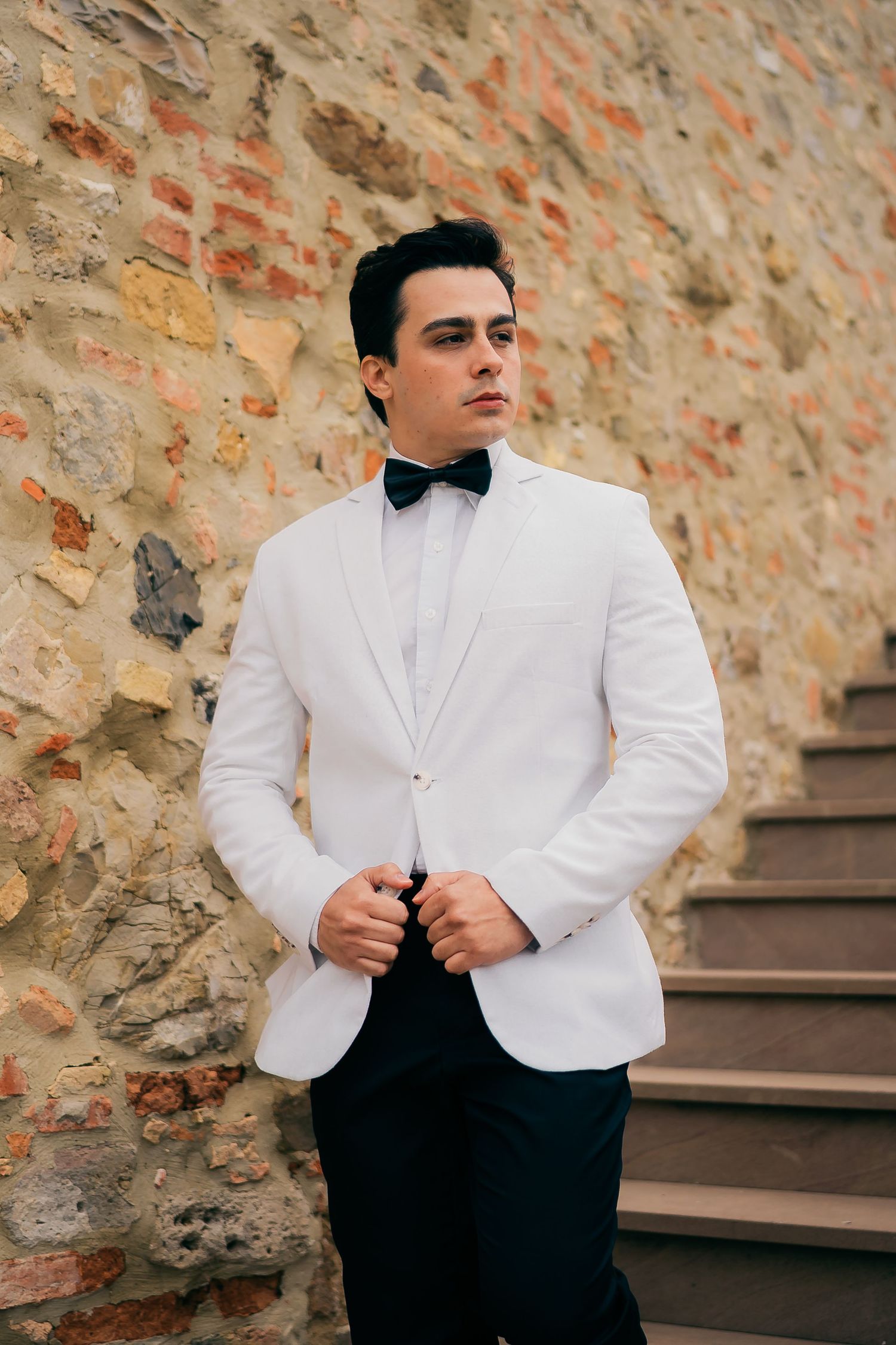 A formal portrait of someone wearing a classic white dinner jacket with black bow tie against a rustic stone wall.