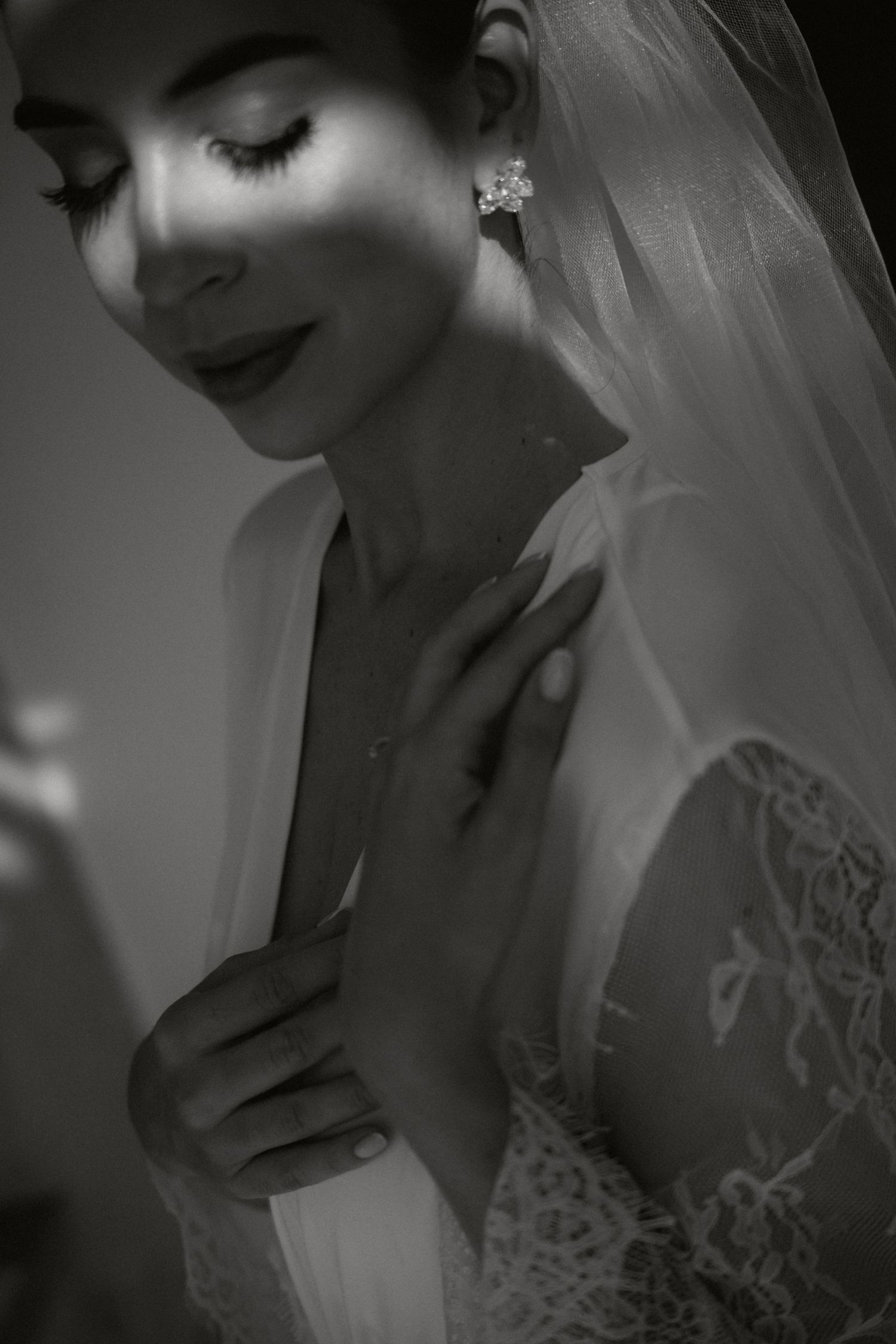 Black and white portrait of a bride in lace gown and veil, wearing crystal earrings, eyes closed, touching her neck.
