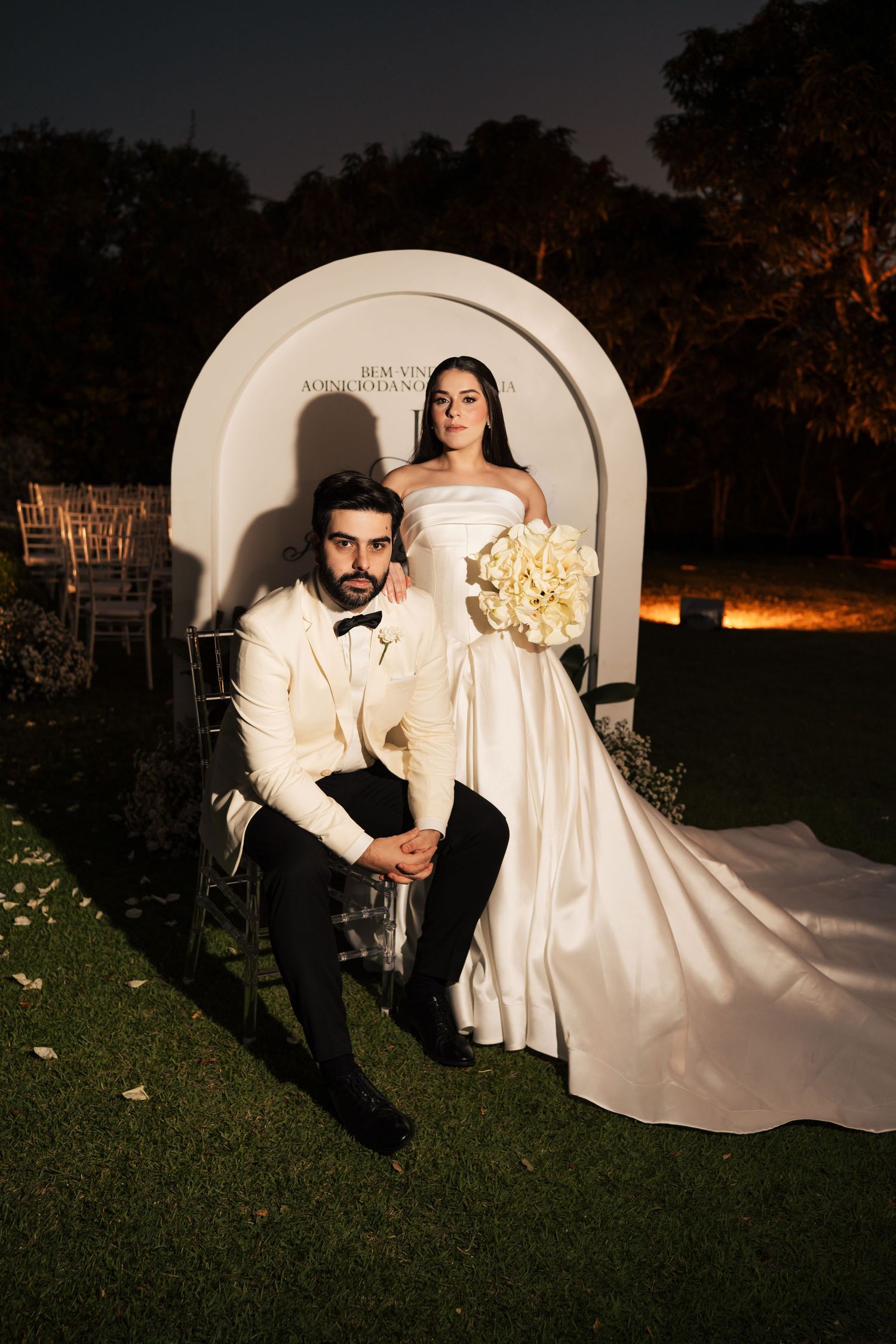 Elegant bride and groom posing at night in front of a white arch, she holds white flowers in a strapless gown.
