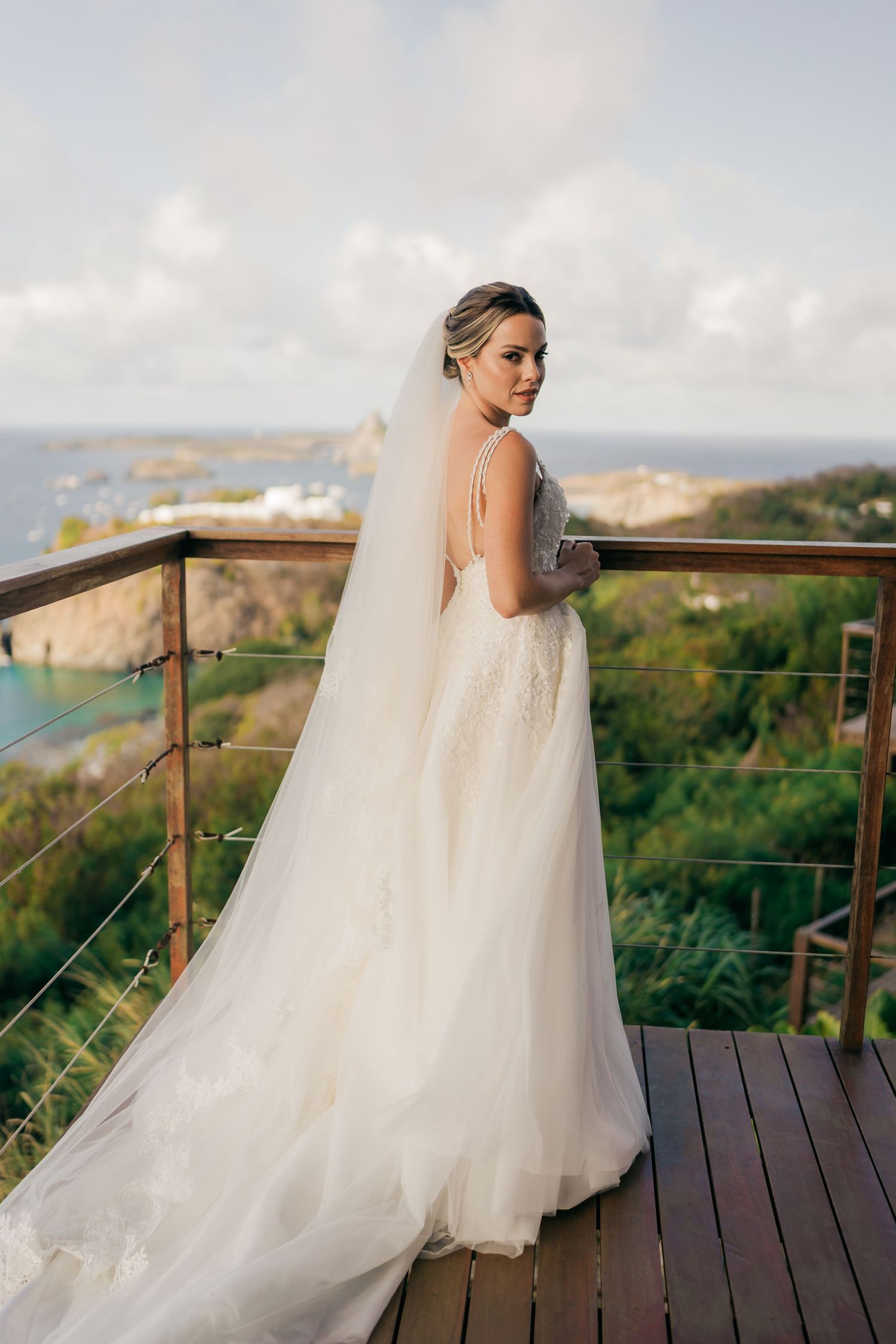 Bride in flowing white gown sitting on wooden deck overlooking scenic mountain landscape.