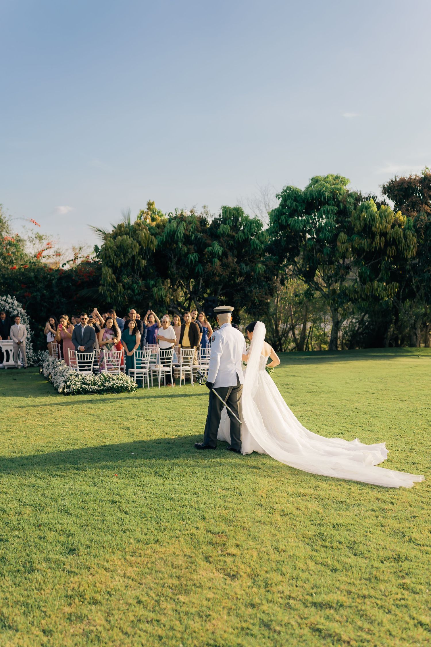 Outdoor wedding ceremony on a lush green lawn with guests seated and a long flowing bridal train.