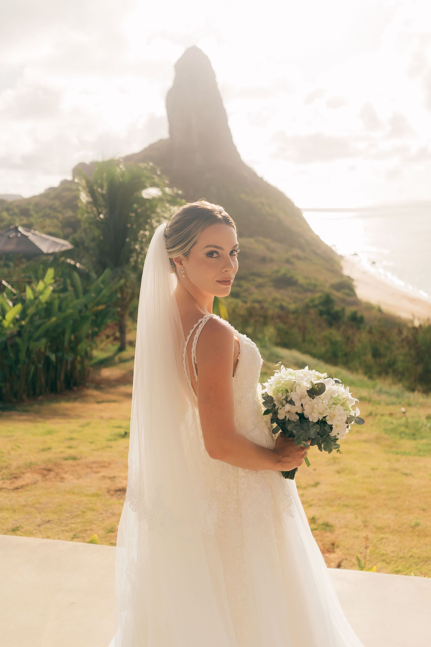 A bride in a flowing white gown holds a bouquet against a scenic mountain backdrop at sunset.