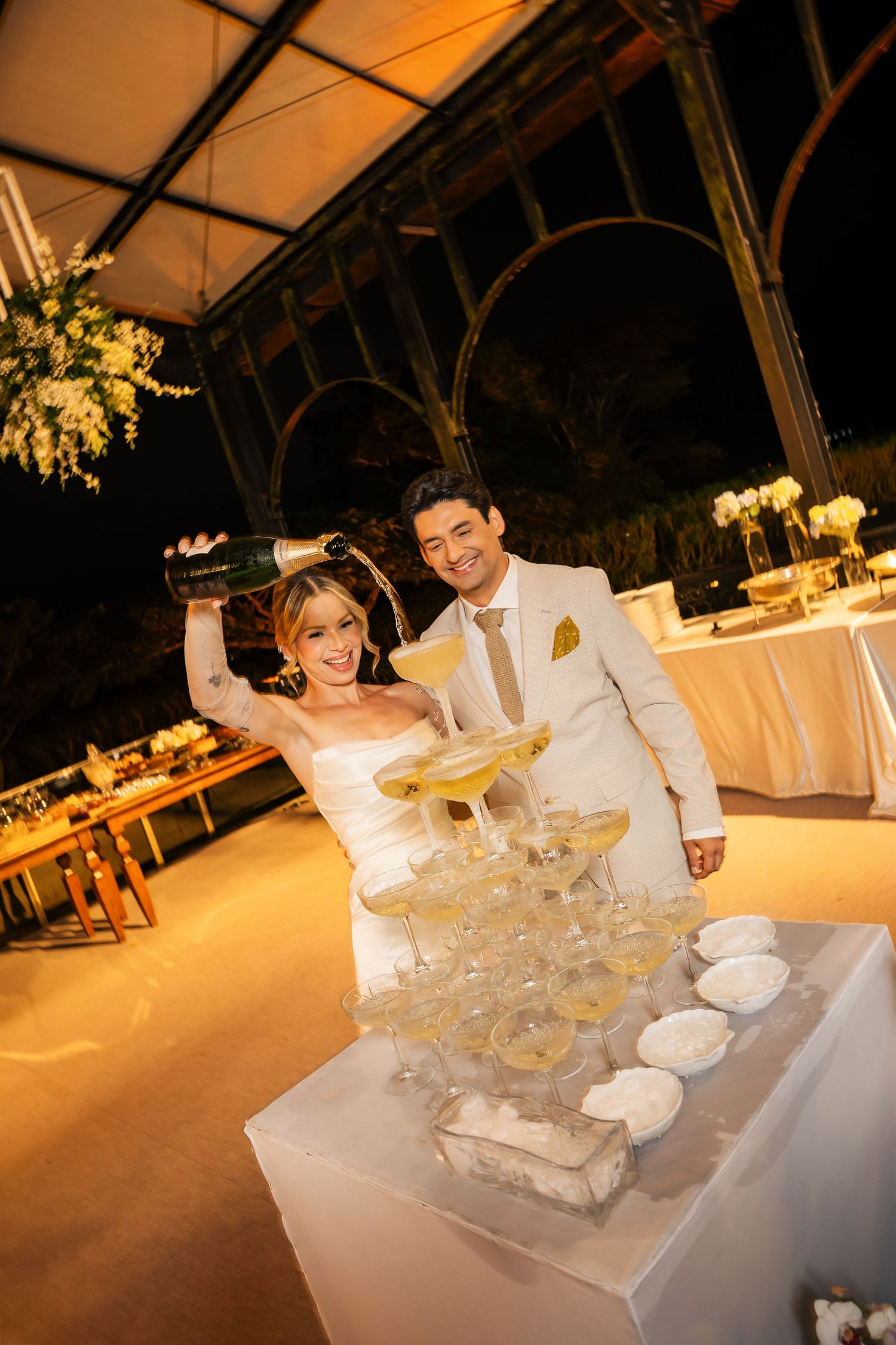 Wedding reception table decorated with elegant white floral centerpieces and glowing ambient lighting in a glass-enclosed venue.