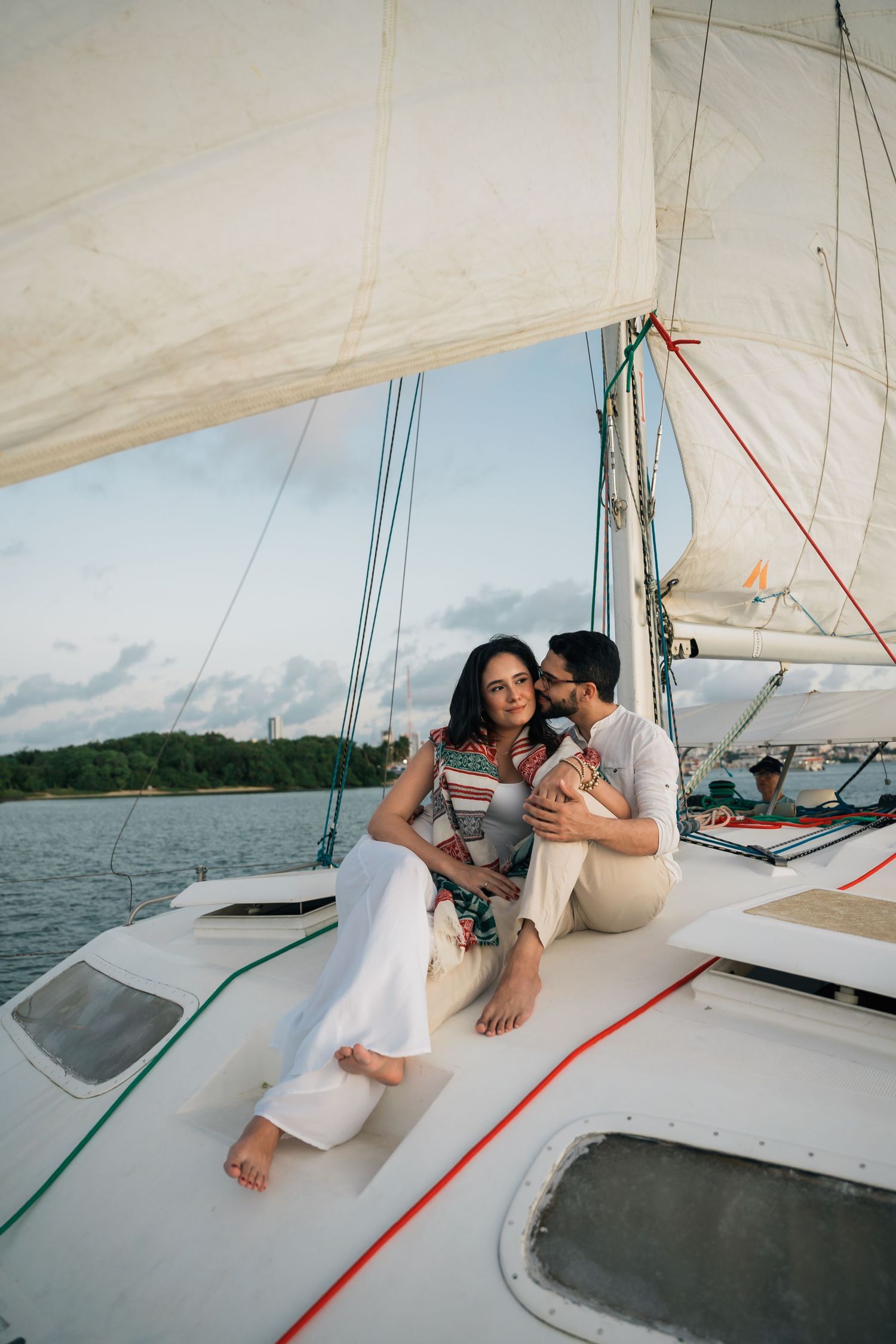 Couple relaxing on a sailboat deck with white sails and blue water in the background.