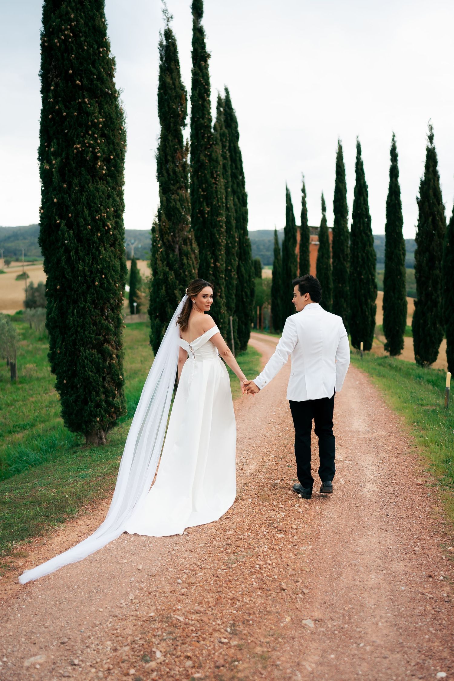 Bride and groom walking hand in hand down a cypress tree-lined gravel path on their wedding day.