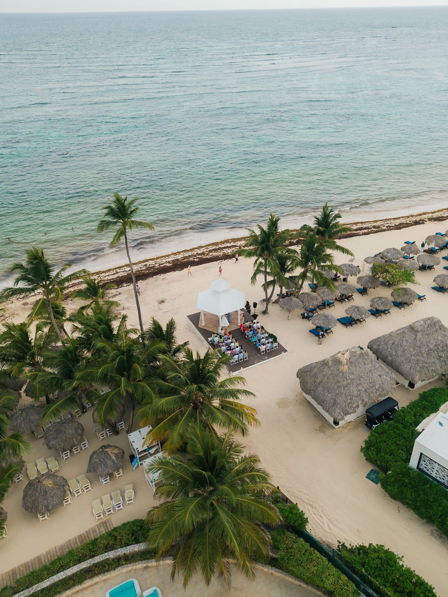Aerial view of a tropical beach with palm trees, loungers, and thatched umbrellas along turquoise waters.