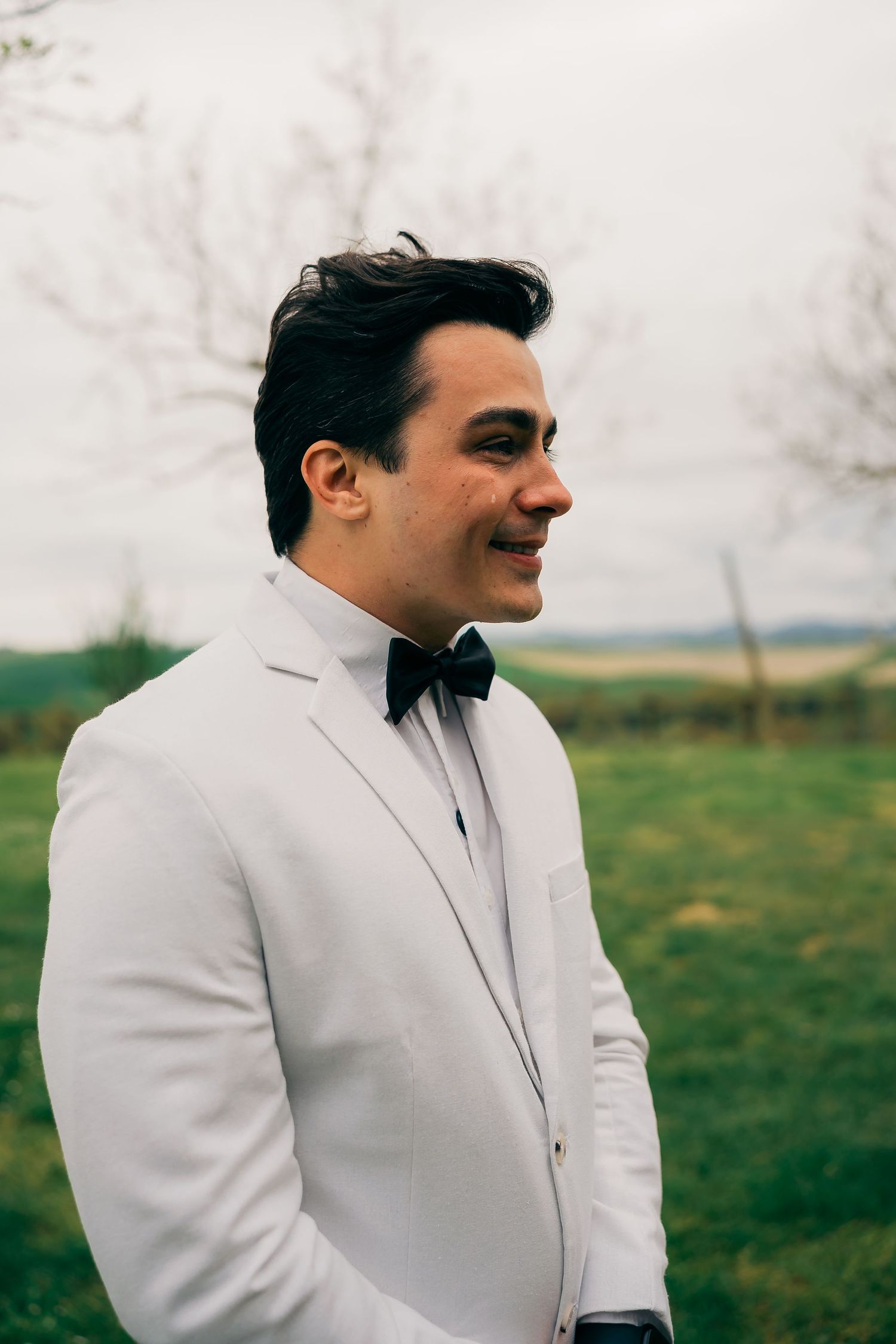 Smiling groom in white tuxedo jacket and black bow tie standing outdoors in a green field on a cloudy day.