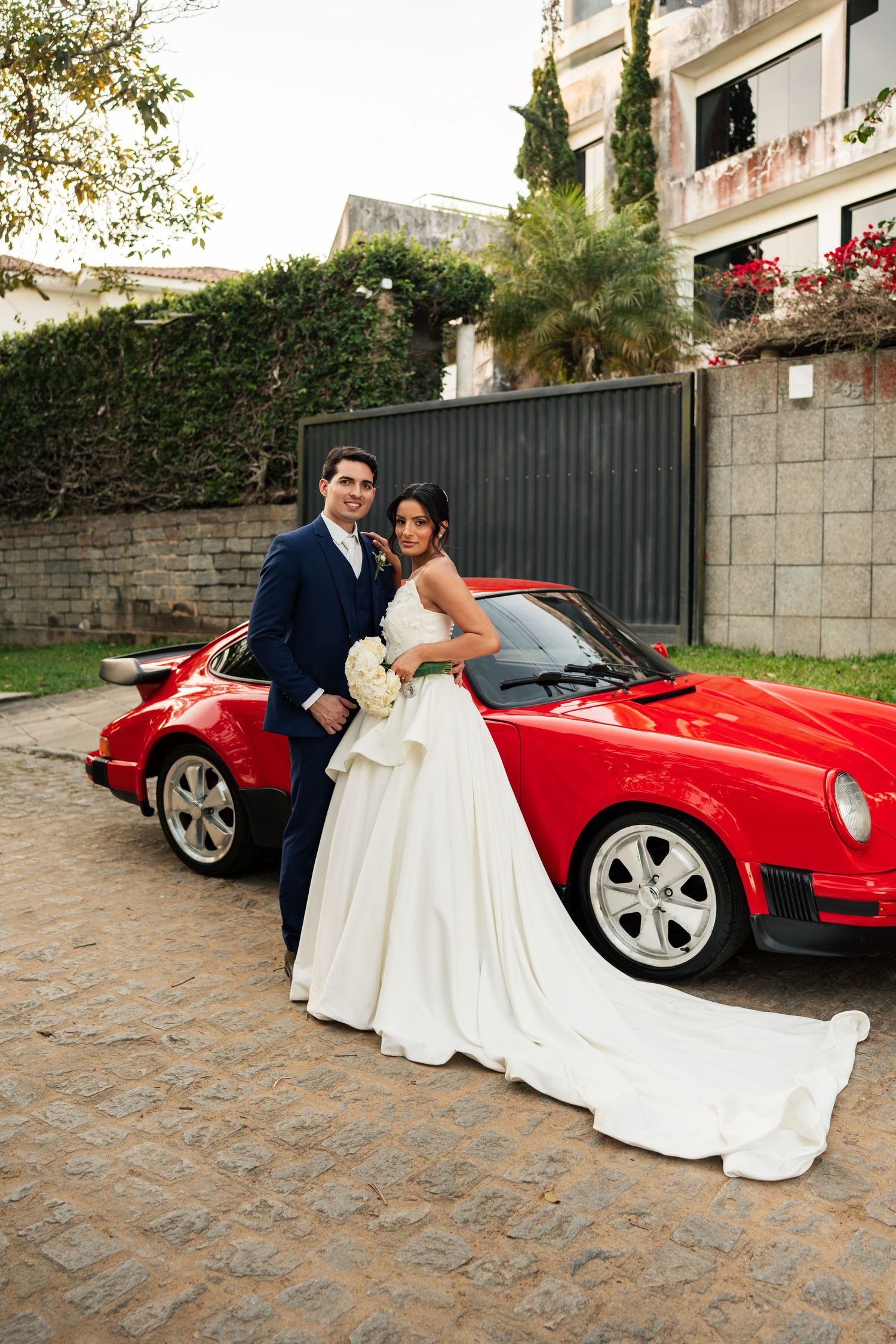 Bride and groom pose beside a classic red Porsche 911 on cobblestone, she in white gown, he in navy suit.