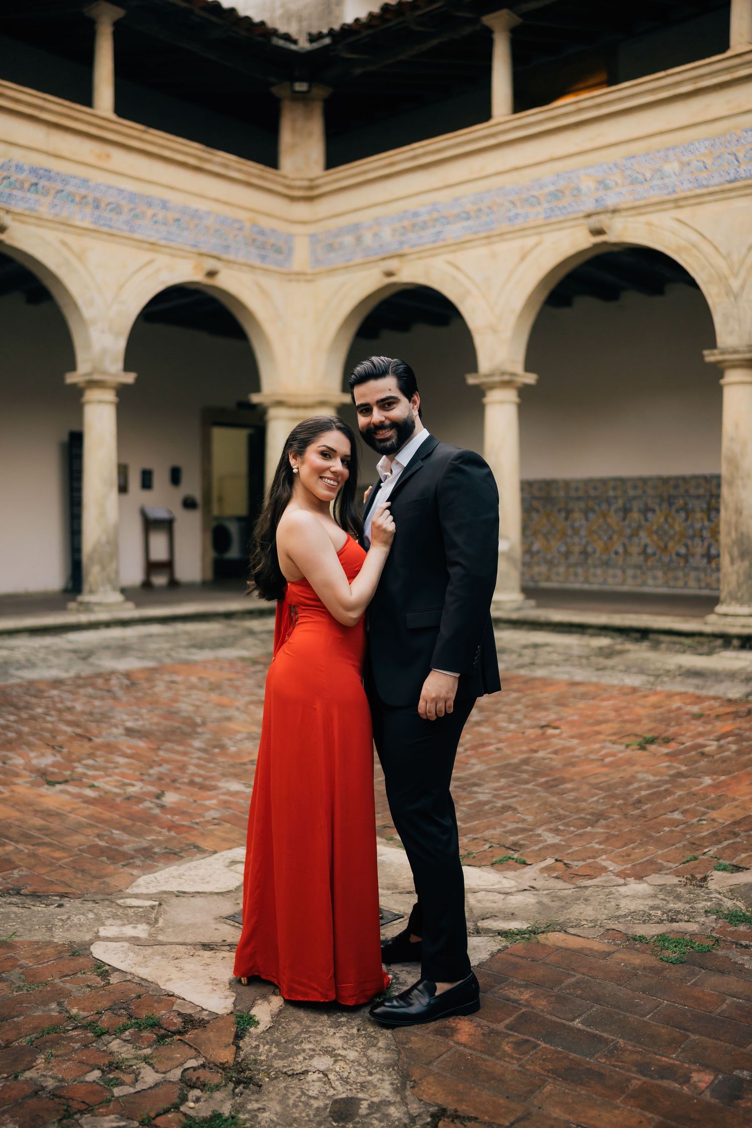 Couple in formal wear embracing in a historic courtyard with arched columns and terracotta floors.