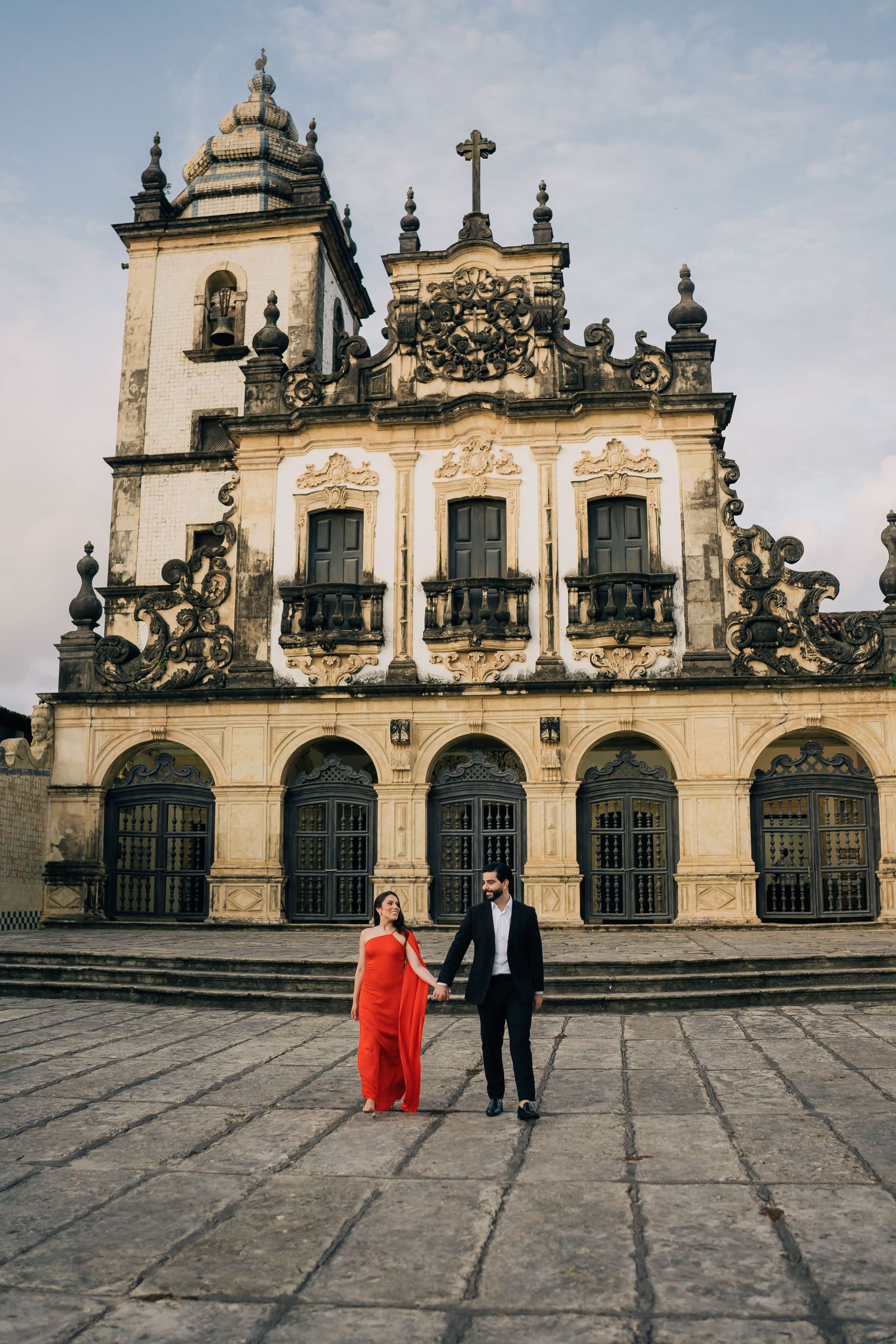 A couple walks in front of an ornate Baroque church facade with decorative spires.