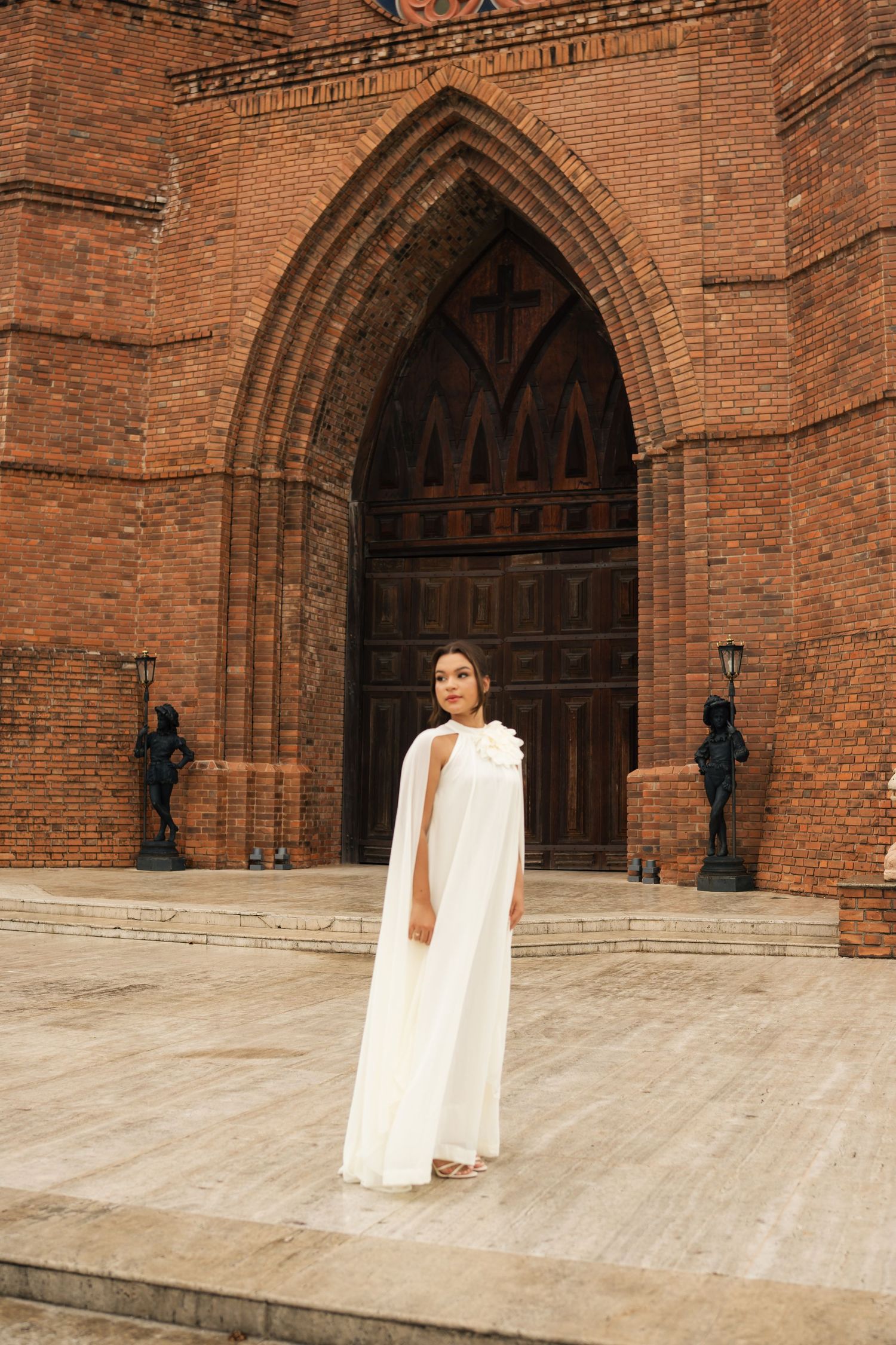 A person in white stands before massive Gothic brick church doors.
