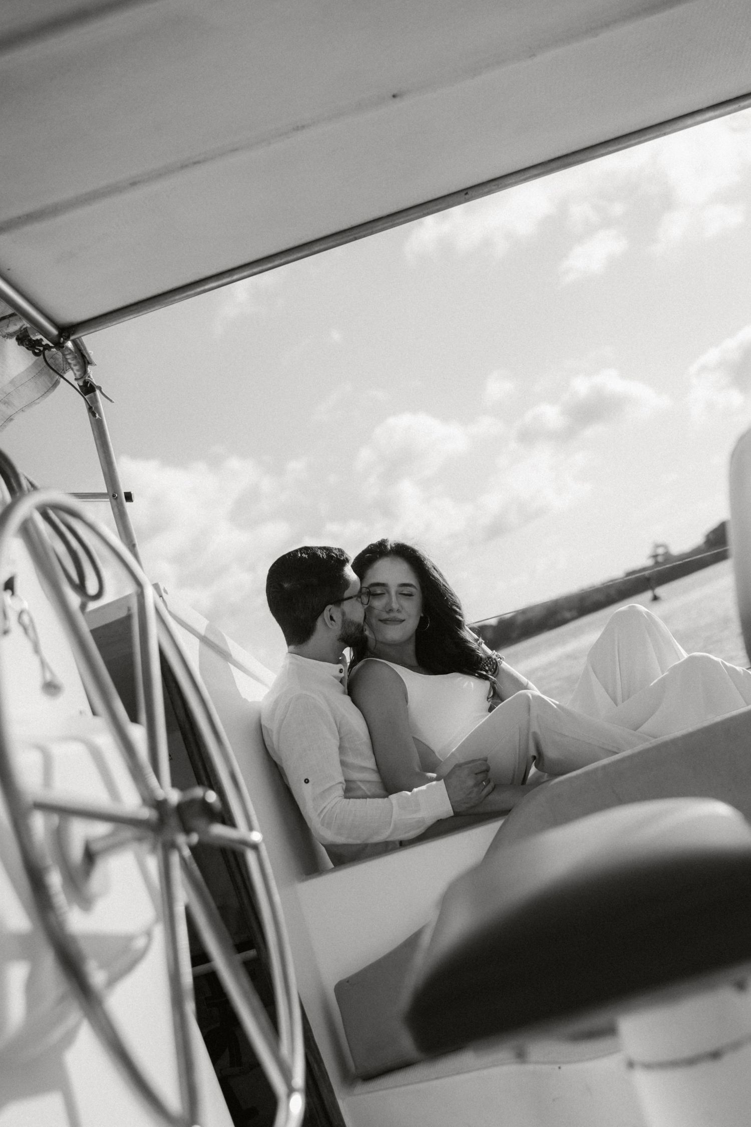 Black and white photo of couple sharing intimate moment on boat deck under cloudy sky.