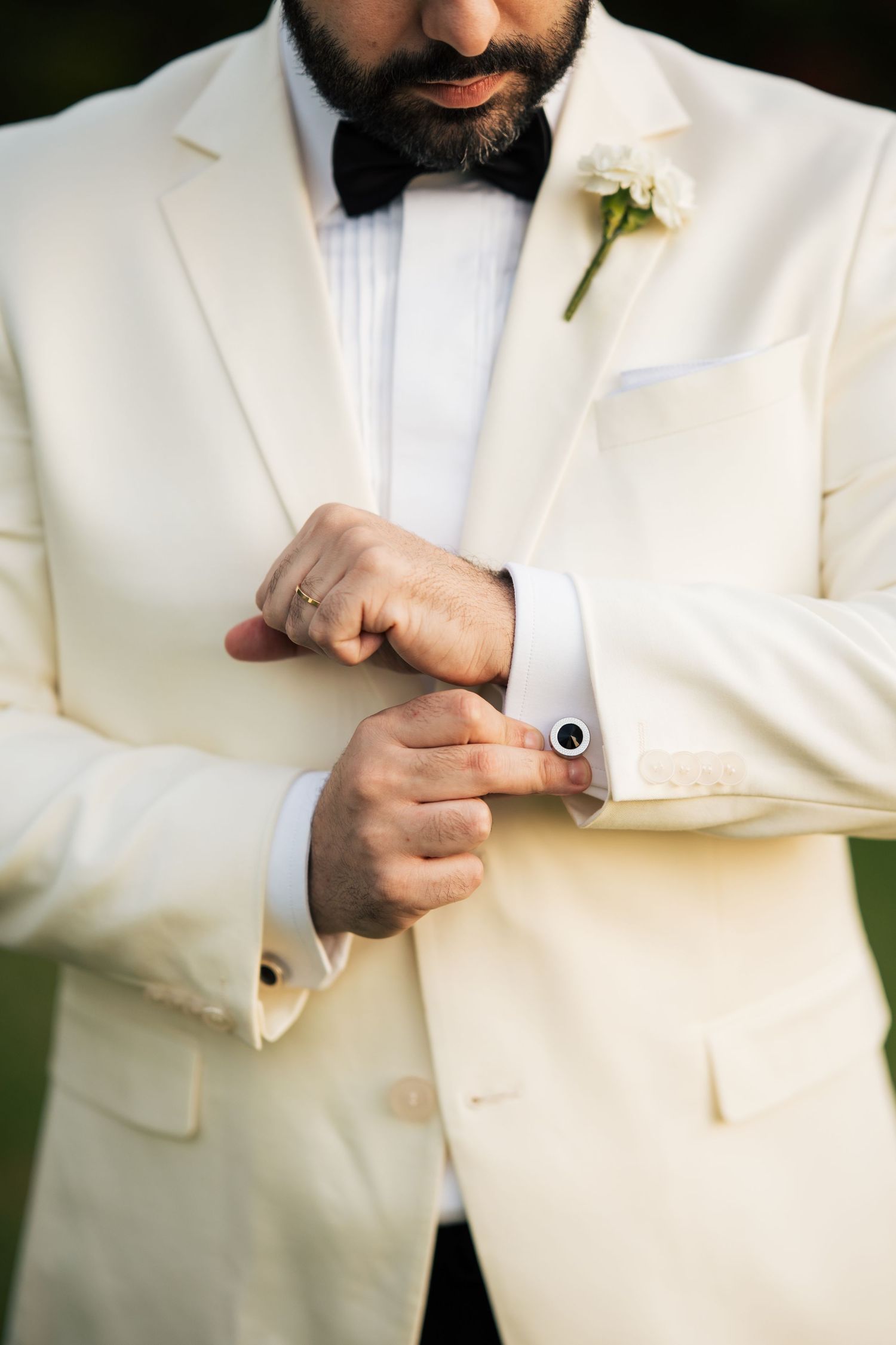 Close-up of groom in cream tuxedo adjusting cufflink with boutonniere on lapel.