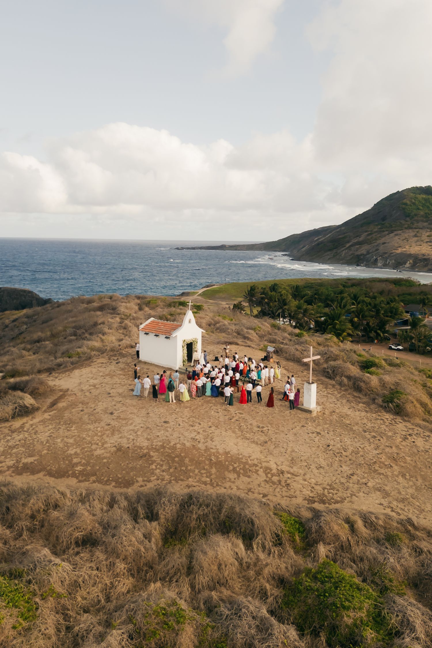 Panoramic coastal landscape with small white chapel and gathering on hilltop overlooking ocean views.